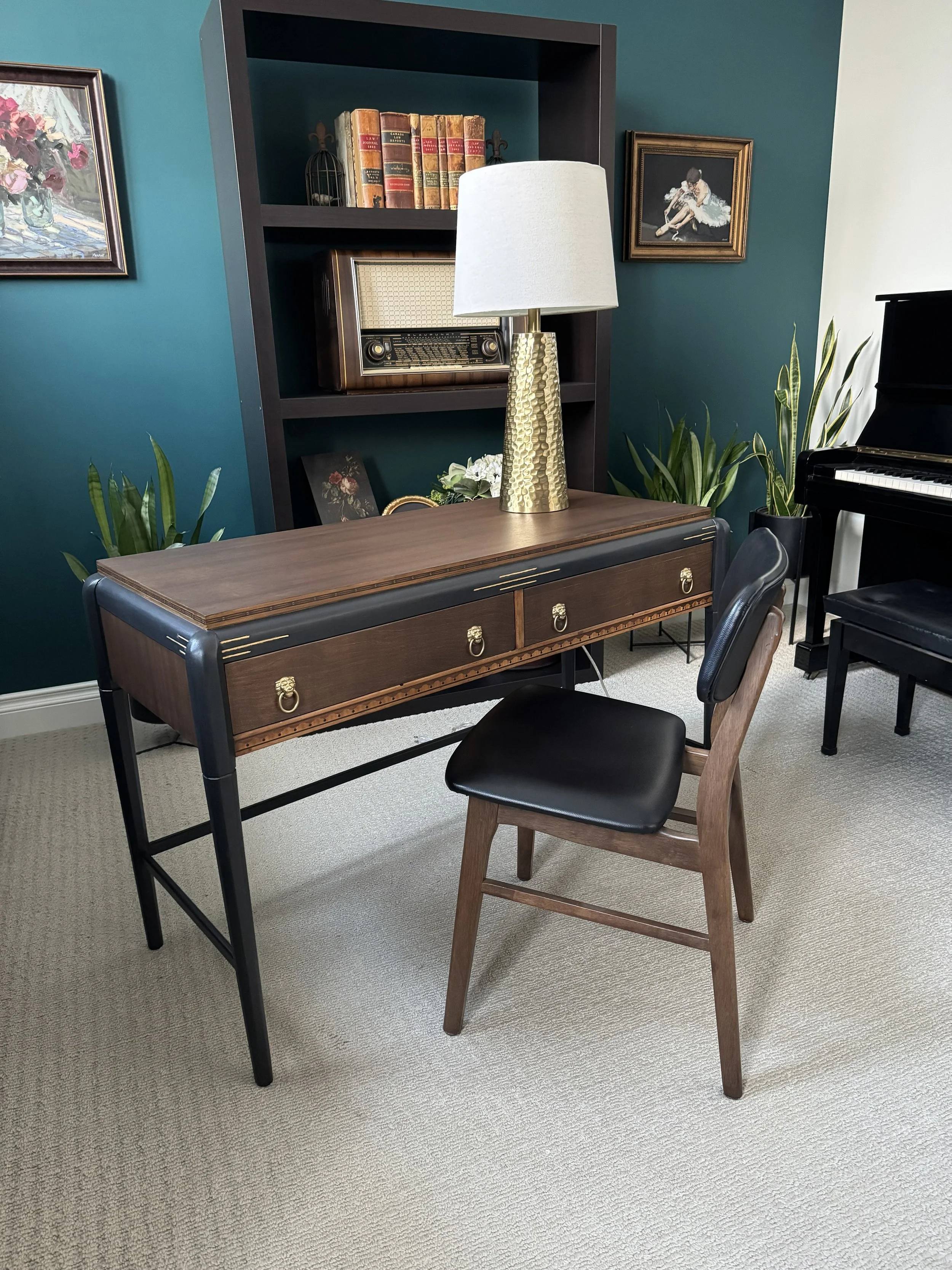 A desk with a gold lamp on top, surrounded by a chair, plants, a bookshelf, and a piano in a room with teal walls.