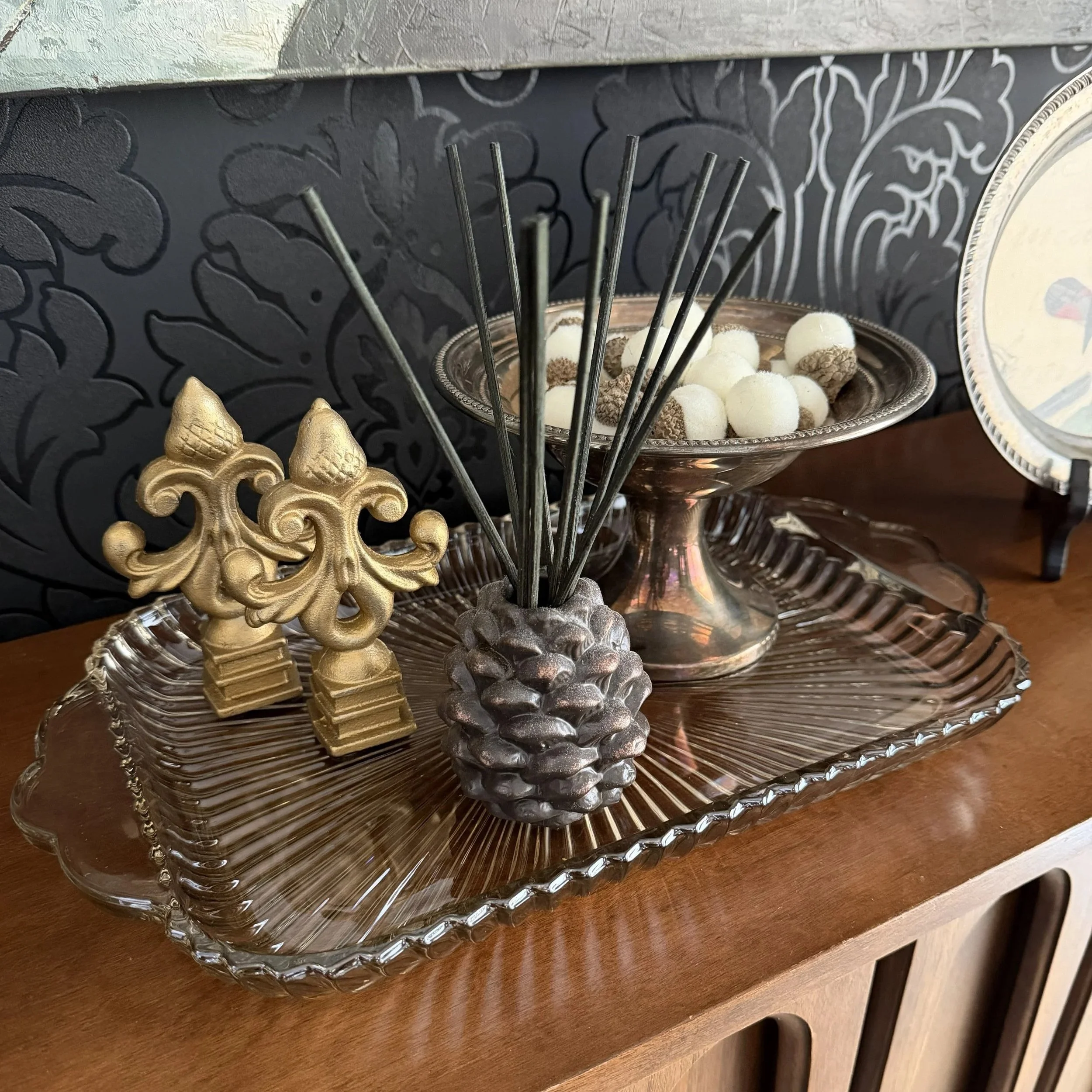 Decorative arrangement on a wooden surface, including two gold ornate pieces, a metallic silver pineapple, a glass tray with a silver dish filled with white and brown candy balls, and a clear glass platter with a parasol design in the background.