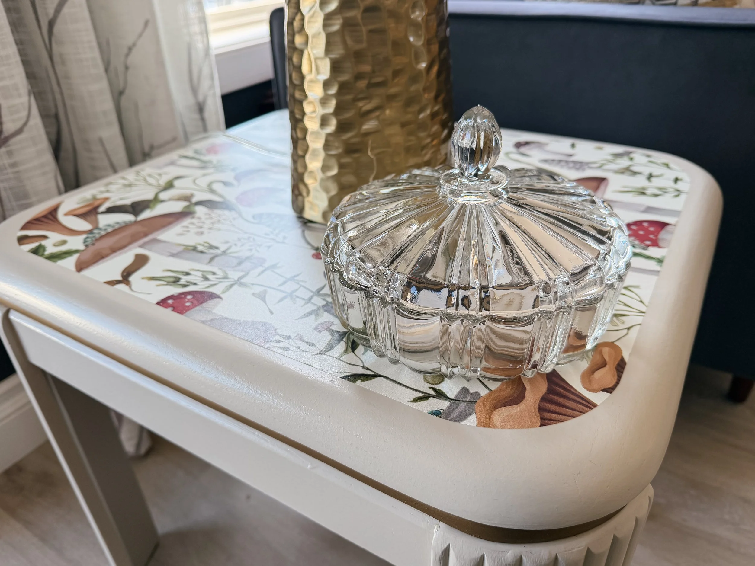 Decorative table with a floral patterned mat, a gold textured vase, a glass jewelry box with a lid, and a beige upholstered chair.