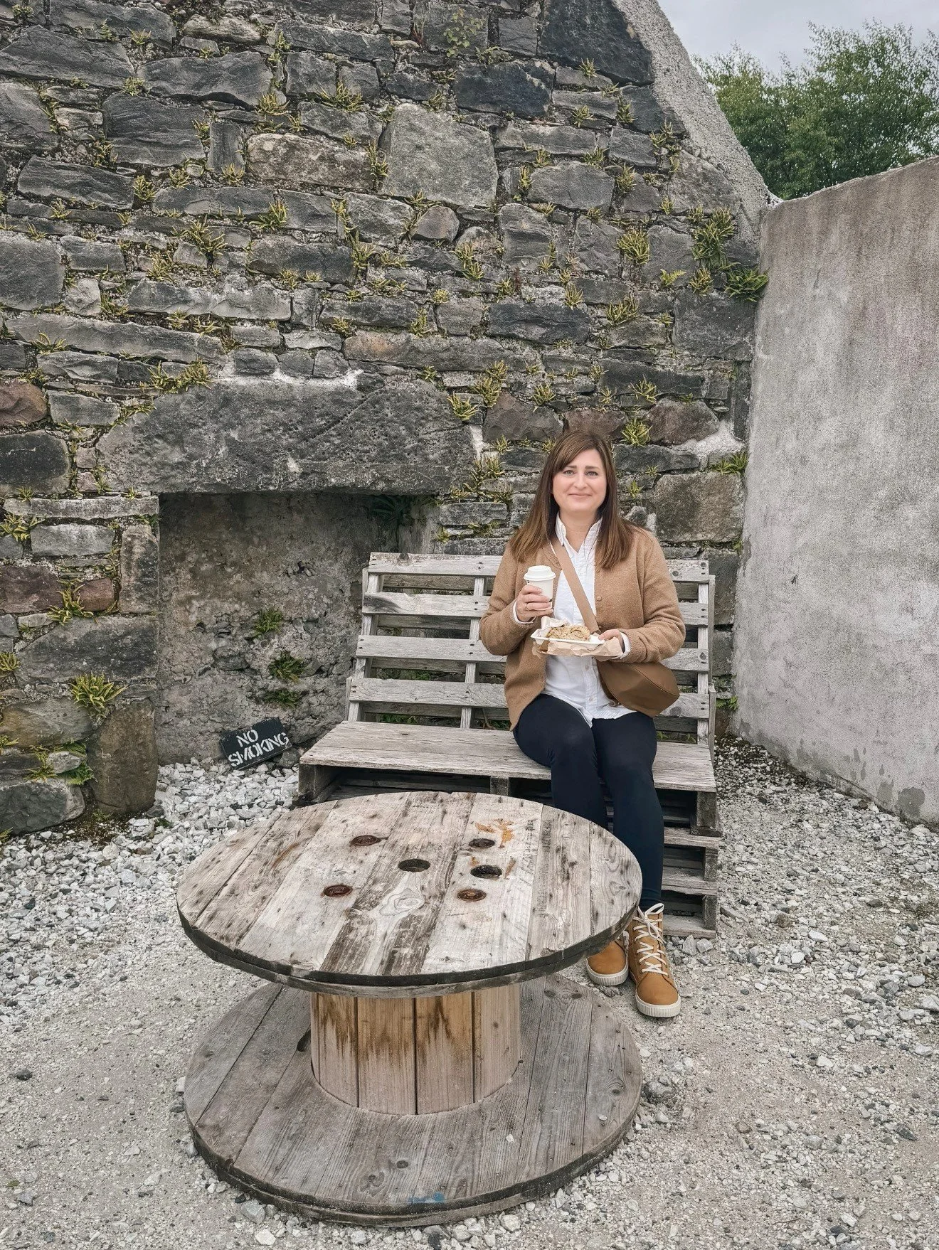 A woman sitting on a wooden bench outdoors, holding a to-go coffee cup and a food item, with a rustic stone wall and a small 'No Smoking' sign behind her.