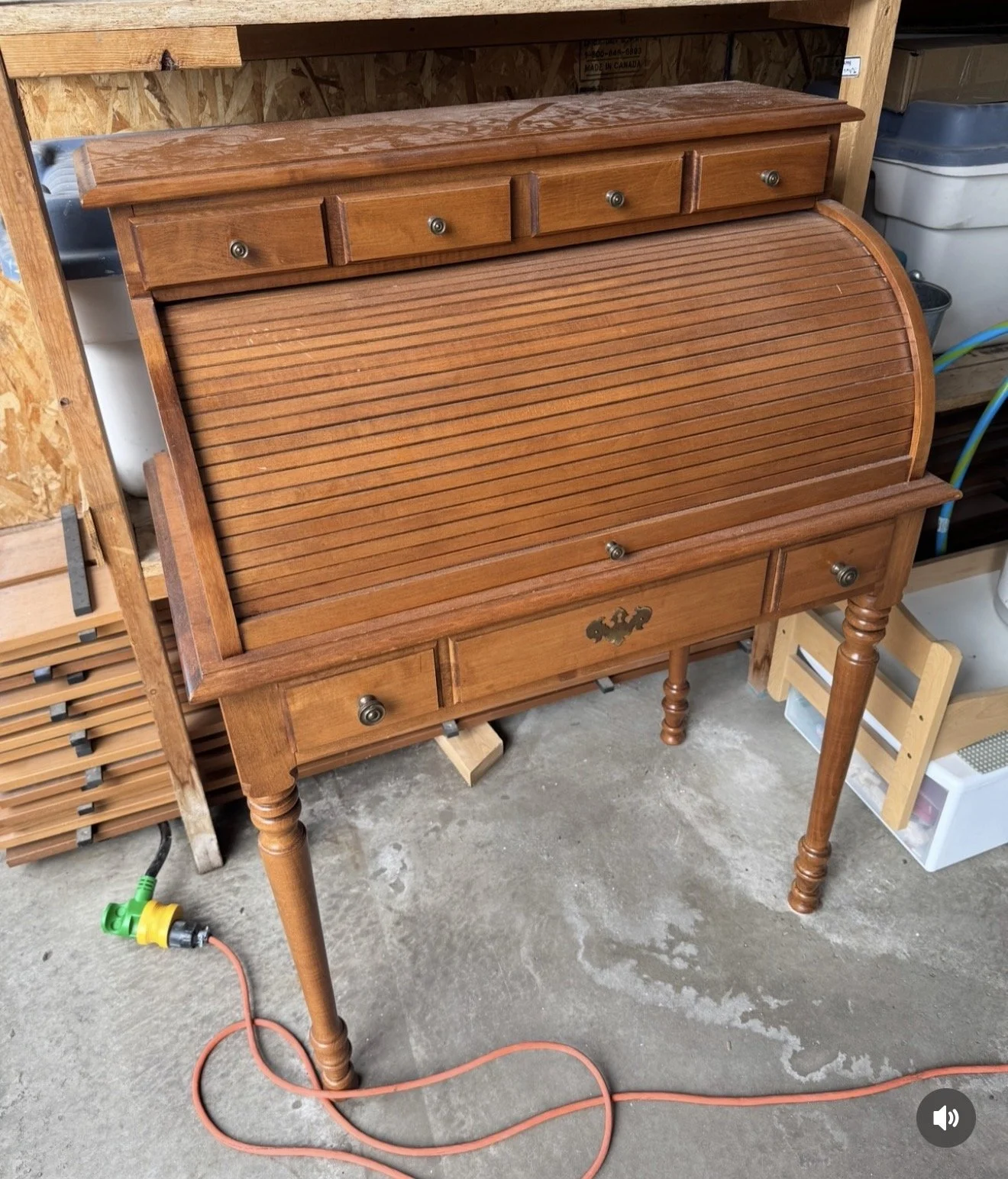 A vintage wooden secretary desk with a roll-top cover, multiple small drawers, and decorative hardware, placed on a garage concrete floor.