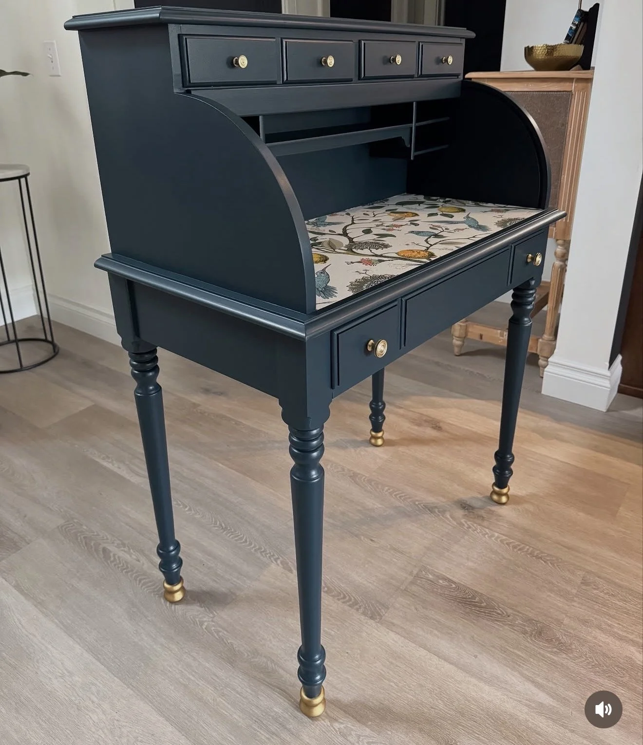 A vintage writing desk painted dark blue with gold knobs on drawers and gold feet, placed on a light wooden floor in a room.
