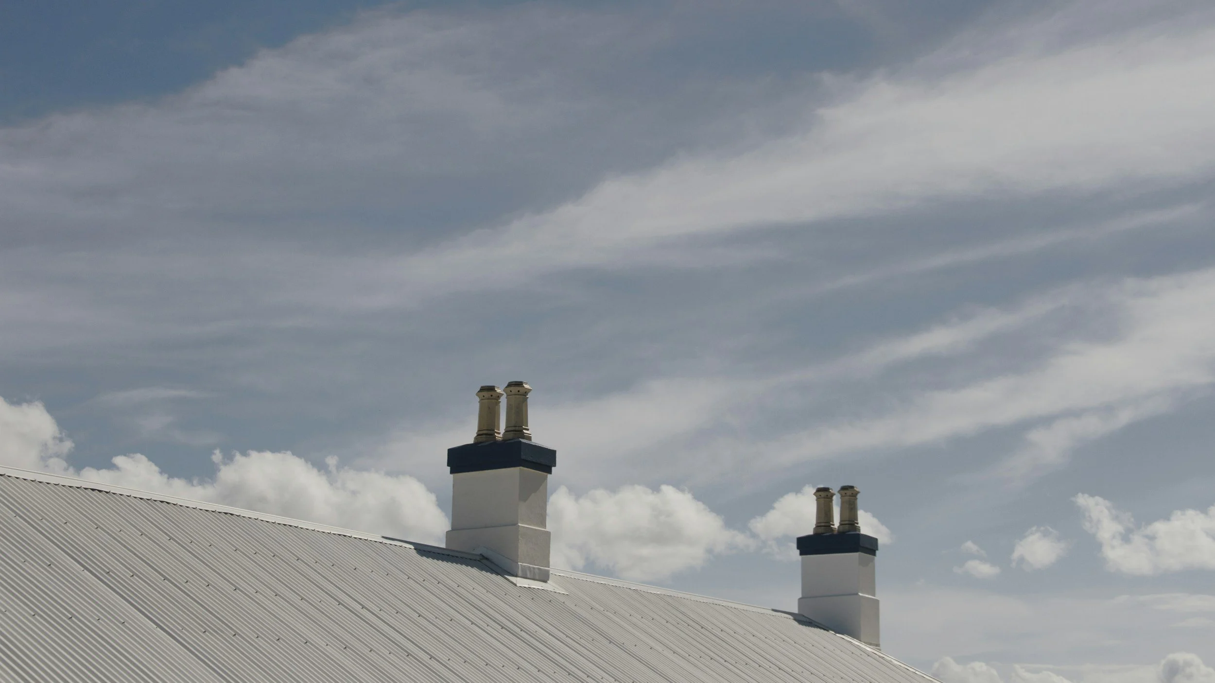 Two white chimneys with beige caps on a corrugated metal roof against a partly cloudy sky.
