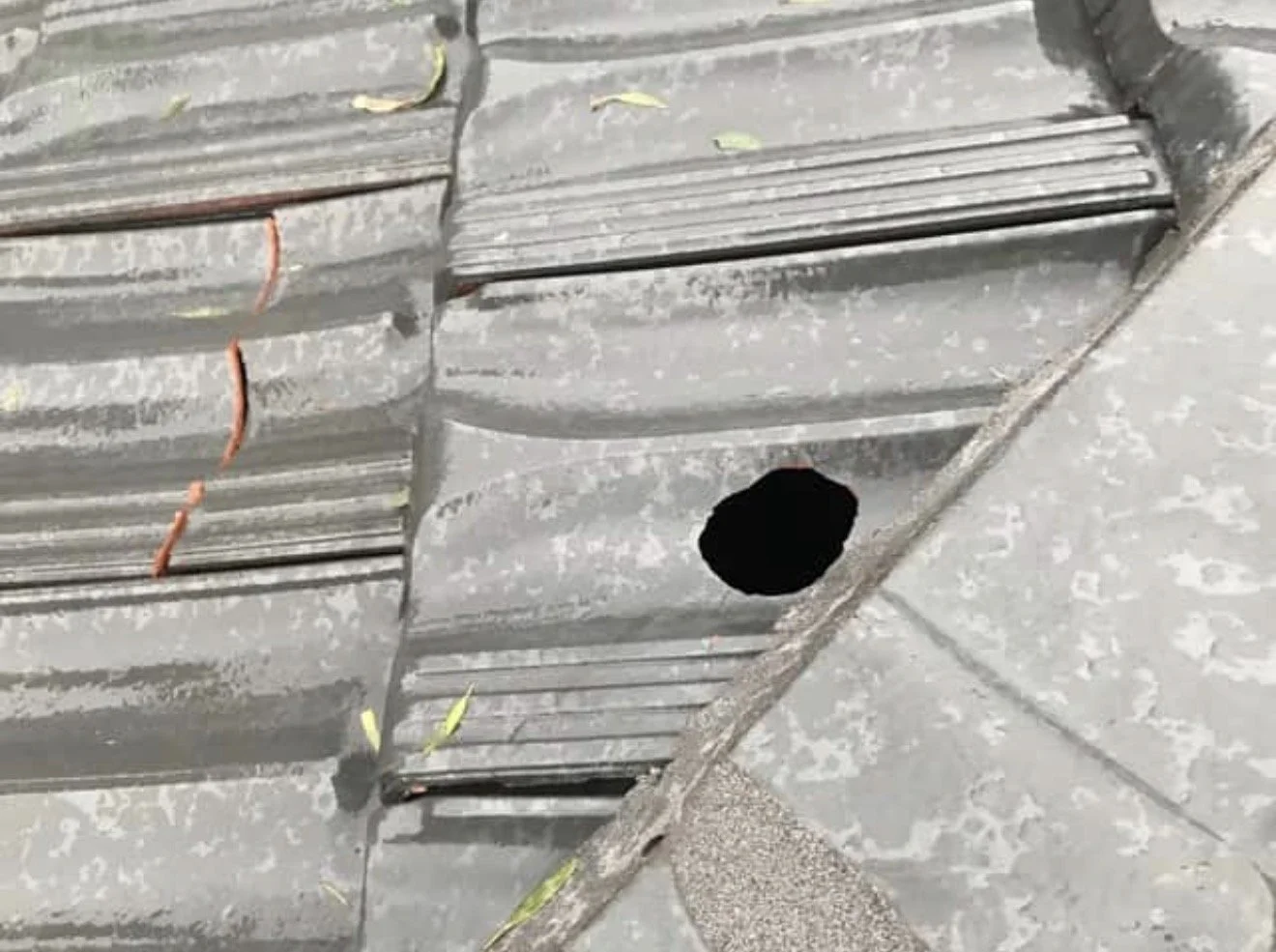 A sidewalk with a square-shaped hole in the ground and a metal grate or cover nearby, with some yellow and orange leaf-like objects scattered on the surface.