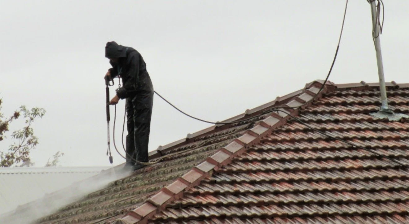 A person wearing a black raincoat and pants, with the hood up, standing on a tiled roof, using a spray tool connected to a hose, likely for cleaning or treating the roof, with an overcast sky in the background.