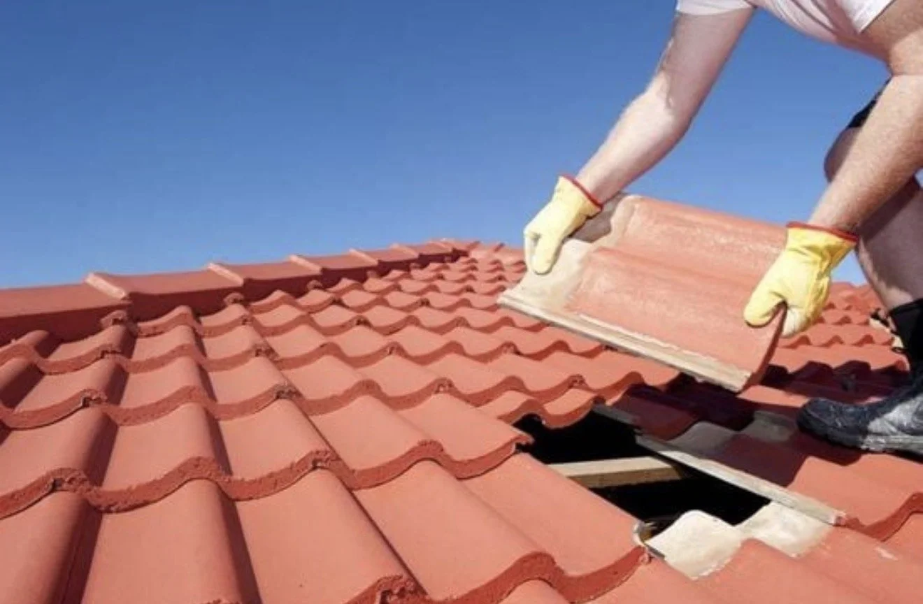 A person installing roof tiles on a red tiled roof, wearing gloves and black shoes, lifting a tile into place under a clear blue sky.