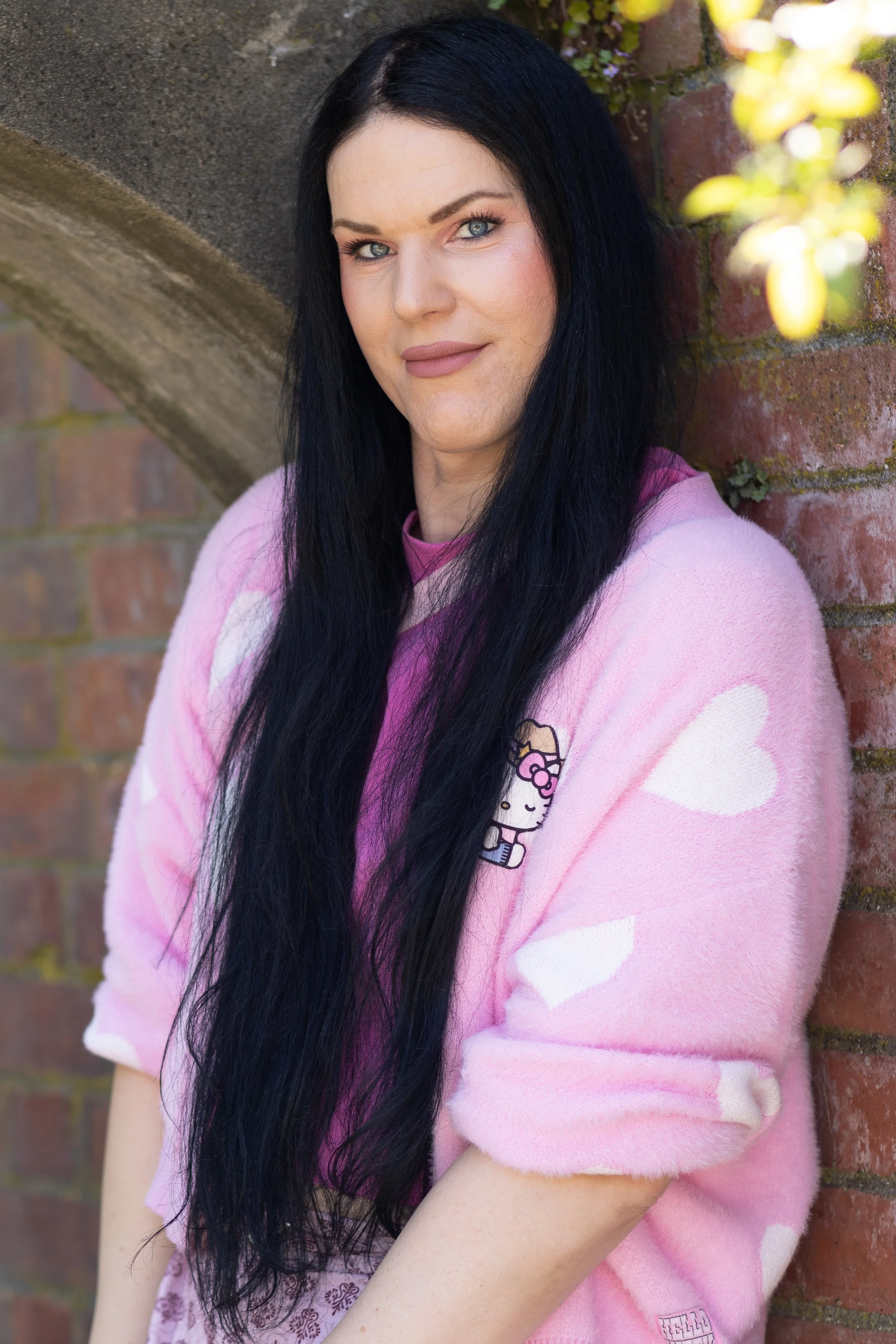 A woman with long black hair and blue eyes leaning against a brick wall, wearing a pink sweater with a Hello Kitty patch and white heart patterns.