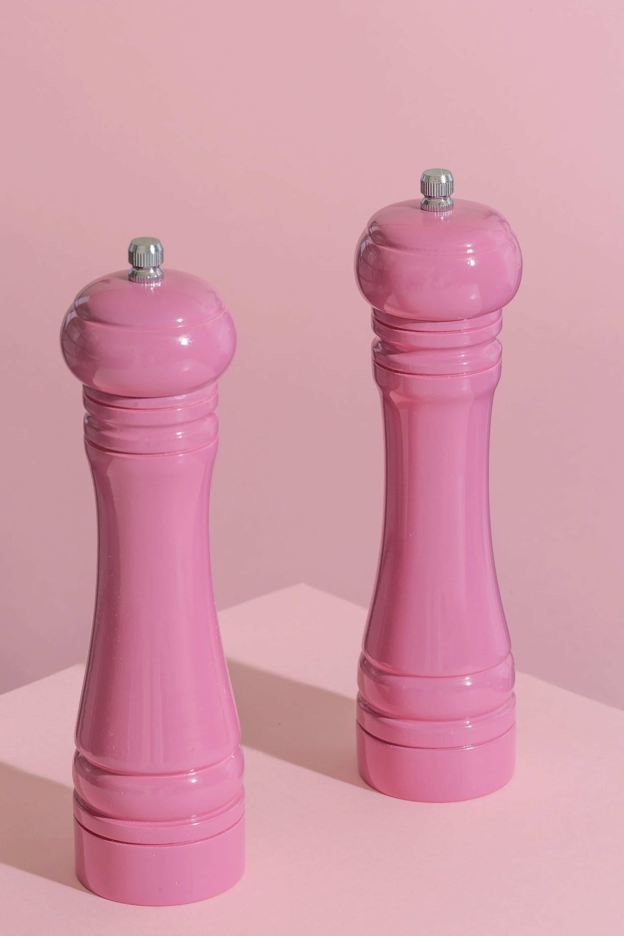 Two pink salt and pepper shakers made of glossy plastic with silver tops, standing on a pink surface against a pink background.