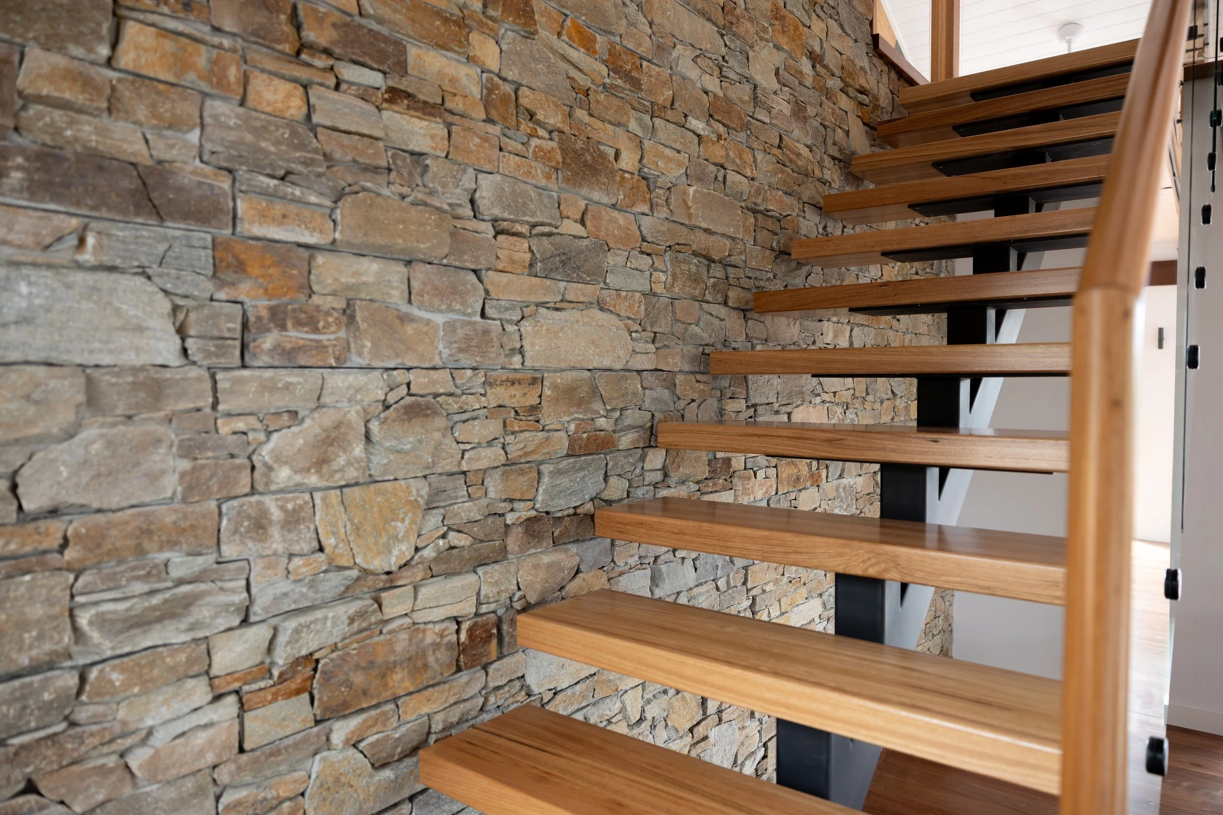 Interior view of a modern blackbutt staircase with wooden steps and black supports, next to a wall made of stacked stone.