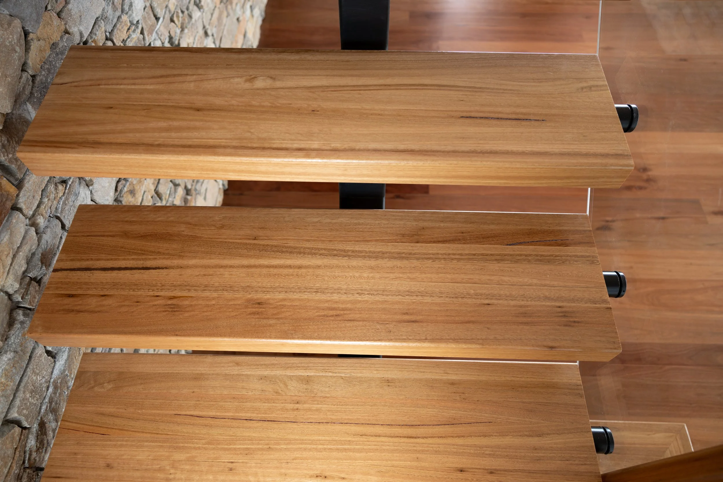 A vertical view of three stairs with a natural finish, positioned against a stone wall and wooden flooring.
