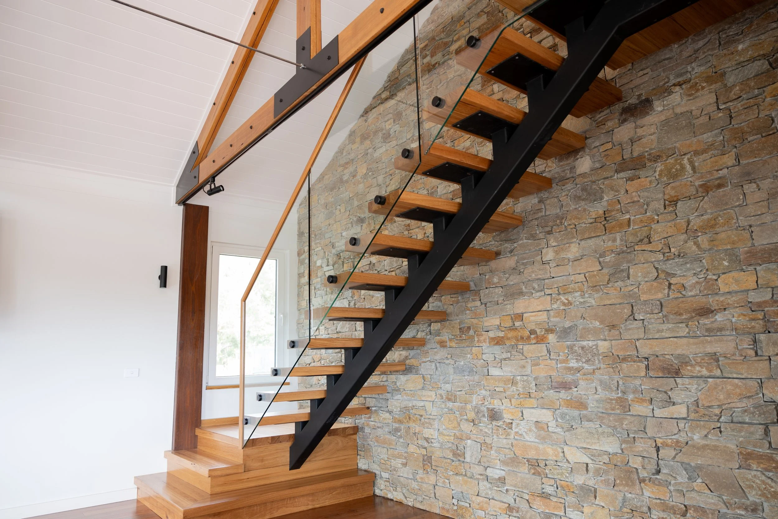 Interior view of a modern blackbutt staircase with wooden steps, a black metal supporting structure, glass railing, and stone wall backdrop in a house with white walls and ceiling.