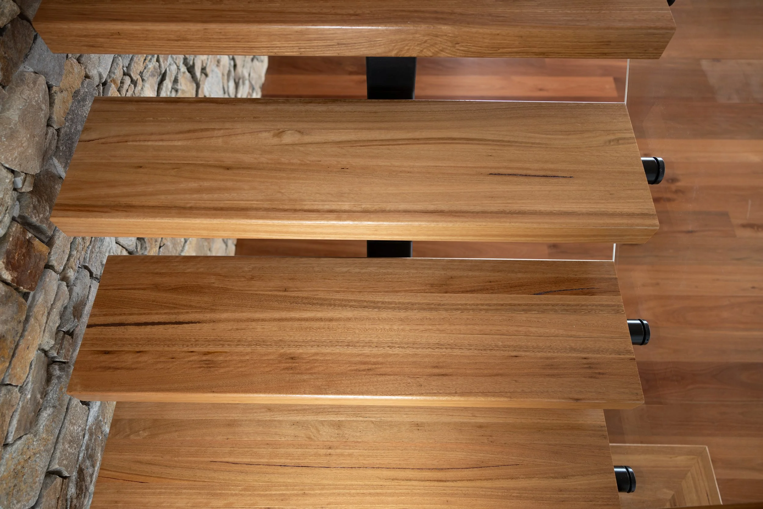 Close-up of wooden stairs attached to a metal frame, against a stone wall on the left side and a hardwood floor in the background.