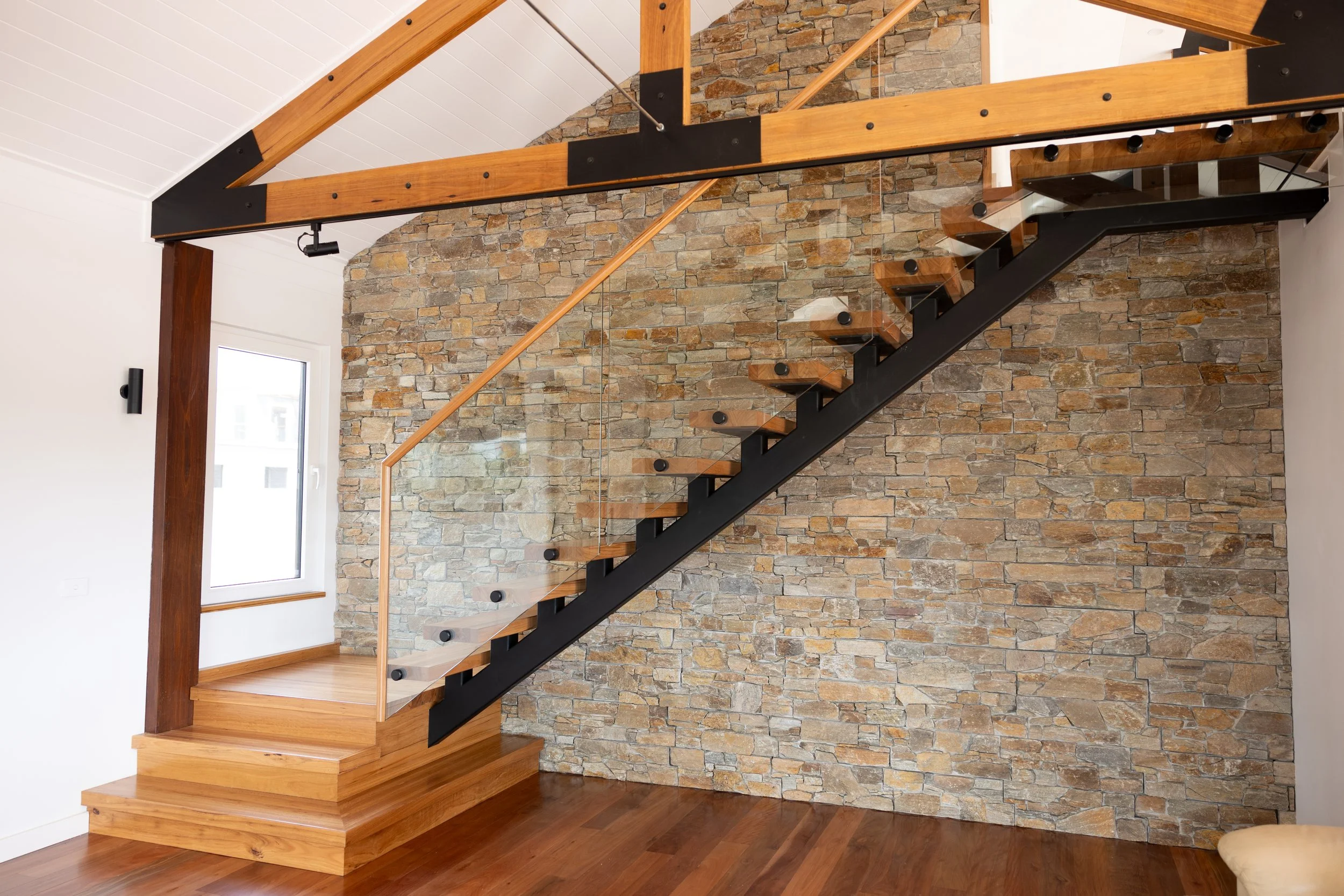 Interior view of a staircase with wooden steps, black metal supports, and glass railing, set against a stone accent wall and white walls.