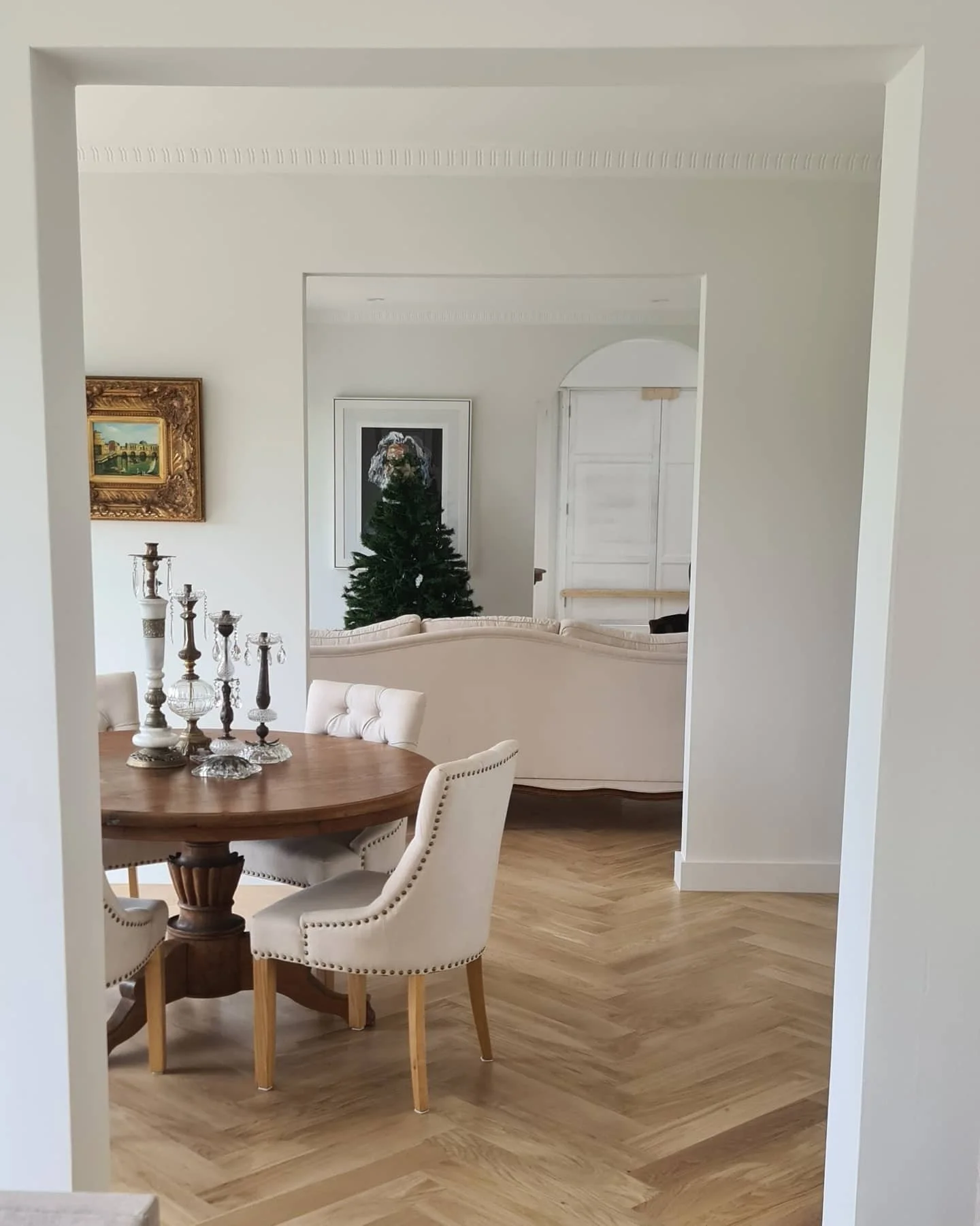 Dining room with round wooden table, white upholstered chairs, candles on table, and a decorated Christmas tree in the background.