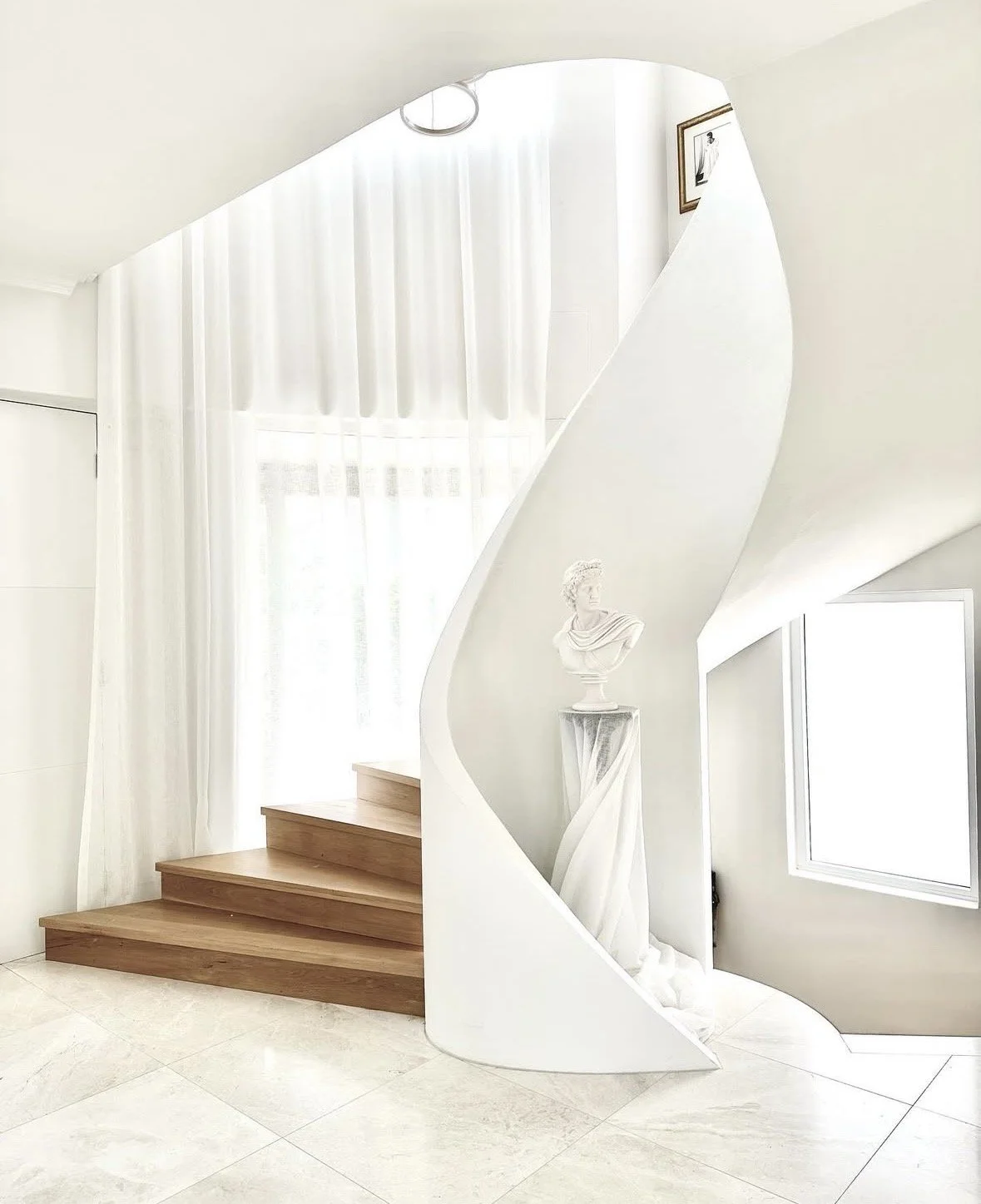 Minimalist staircase with wooden steps, white curved wall, classical bust statue on a pedestal, and framed black-and-white photograph on the wall, with sheer white curtains and a window in the background.