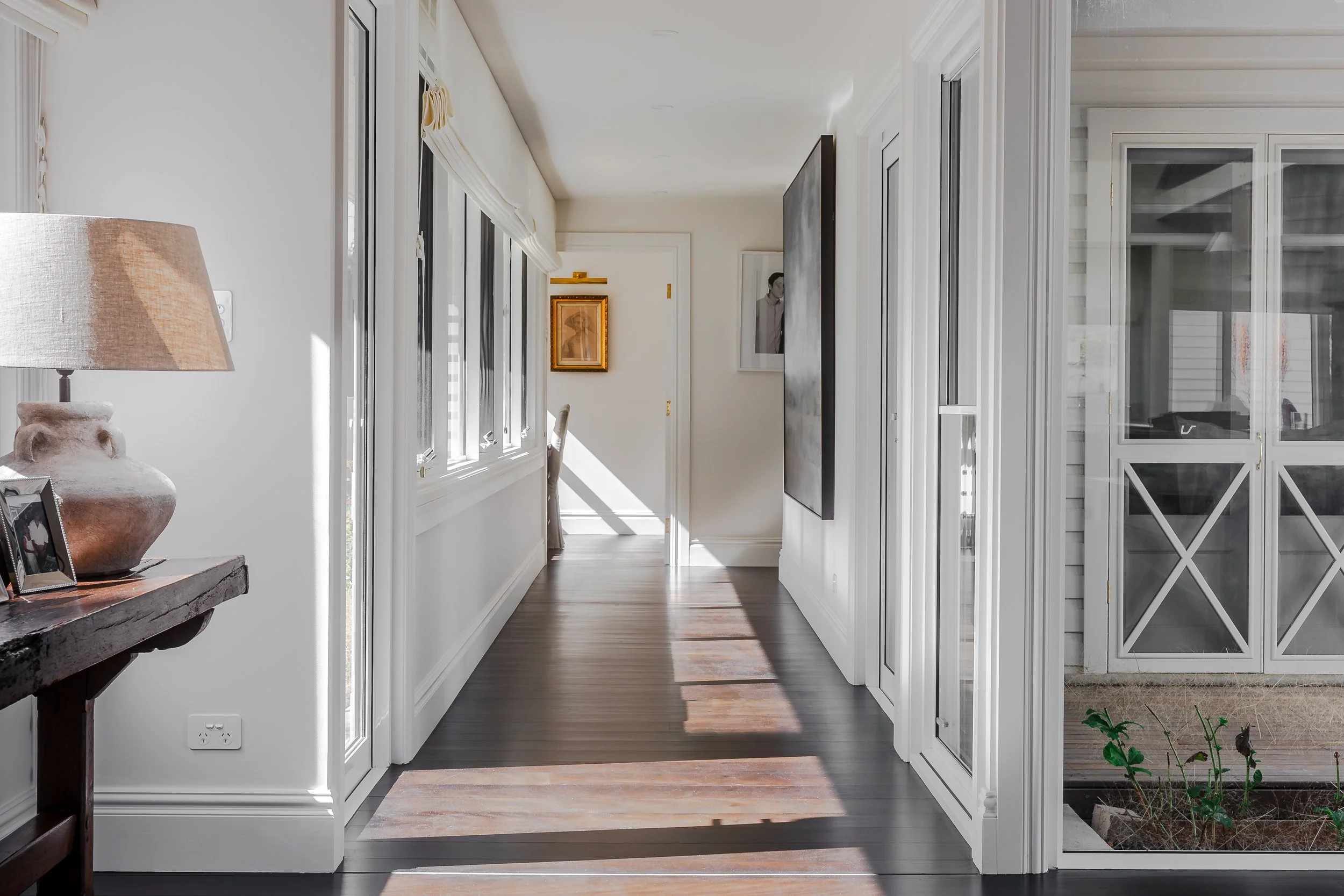 Bright hallway with wooden flooring, white walls, large windows, a door at the far end, and artwork hanging on the walls.