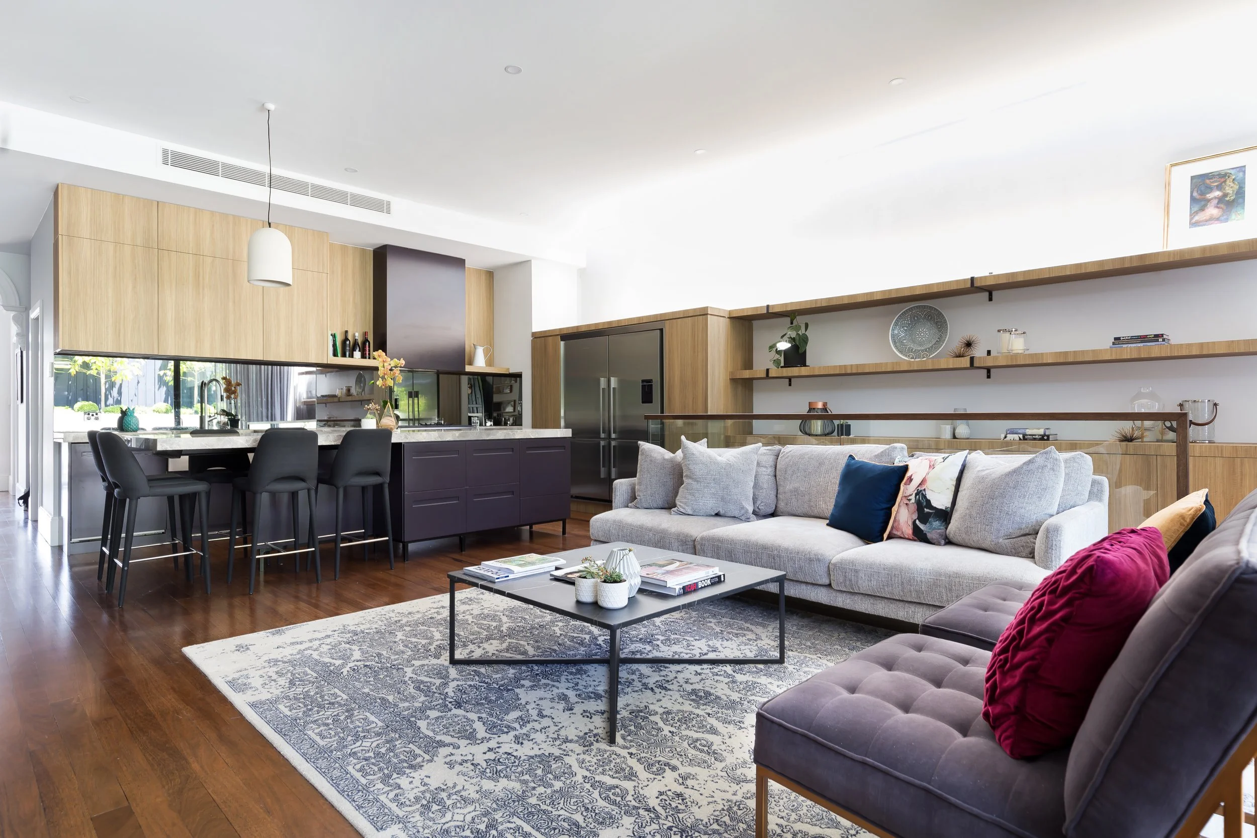Open-concept living room and kitchen with wooden flooring, a large gray sofa with colorful pillows, a patterned area rug, a coffee table with books and plants, and a kitchen with dark lower cabinets, light upper cabinets, and a breakfast bar with four black chairs.