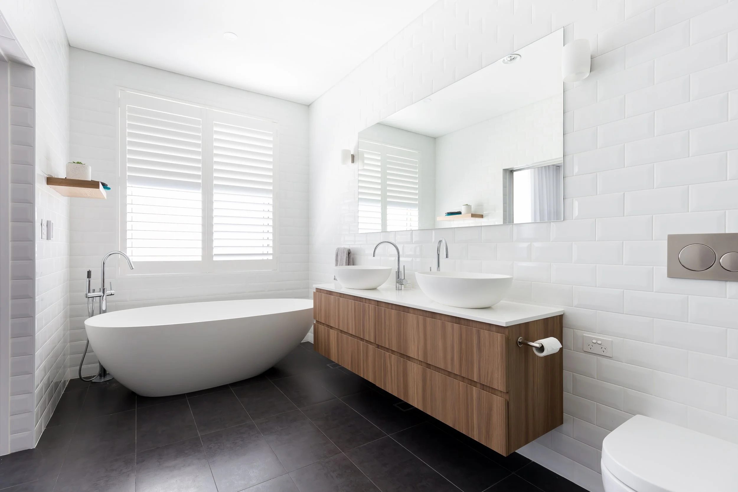 Modern bathroom with white brick walls, a freestanding bathtub, double vessel sinks on a wooden vanity, a large mirror, and black tiled floor, illuminated by natural light from a window.