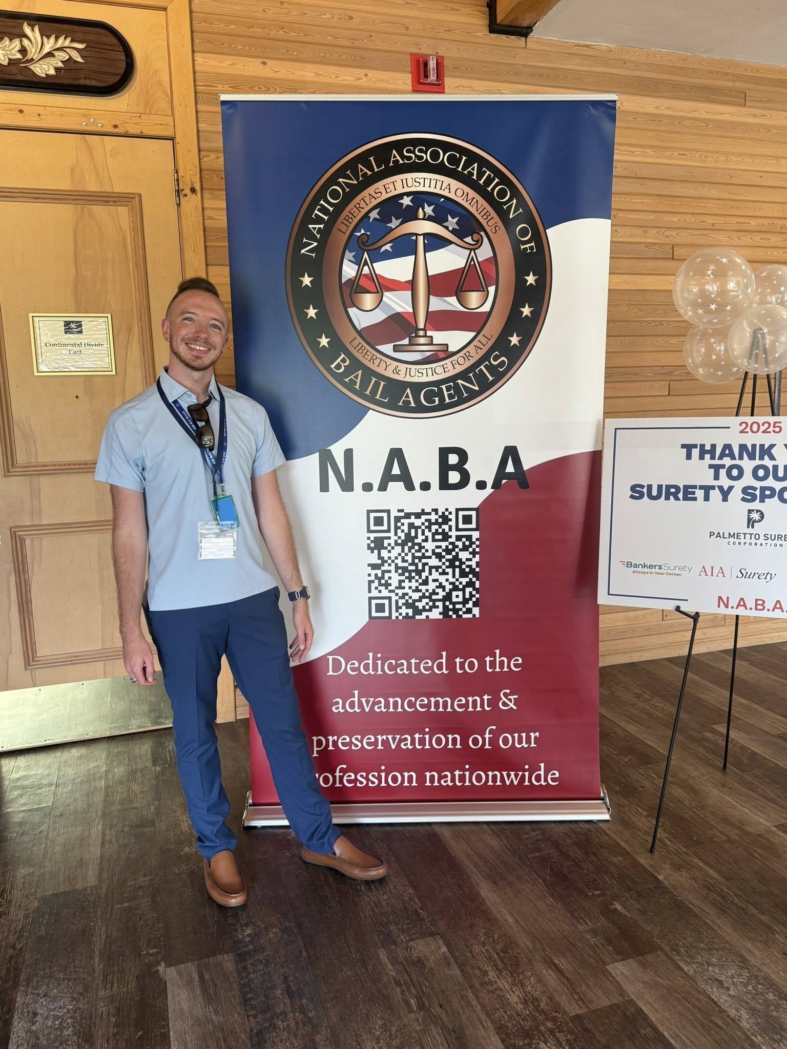 Joshua is smiling and standing next to a large banner for the National Association of Bail Agents (N.A.B.A). The banner features the organization’s logo, a balance scale and American flag design, and text thanking supporters.