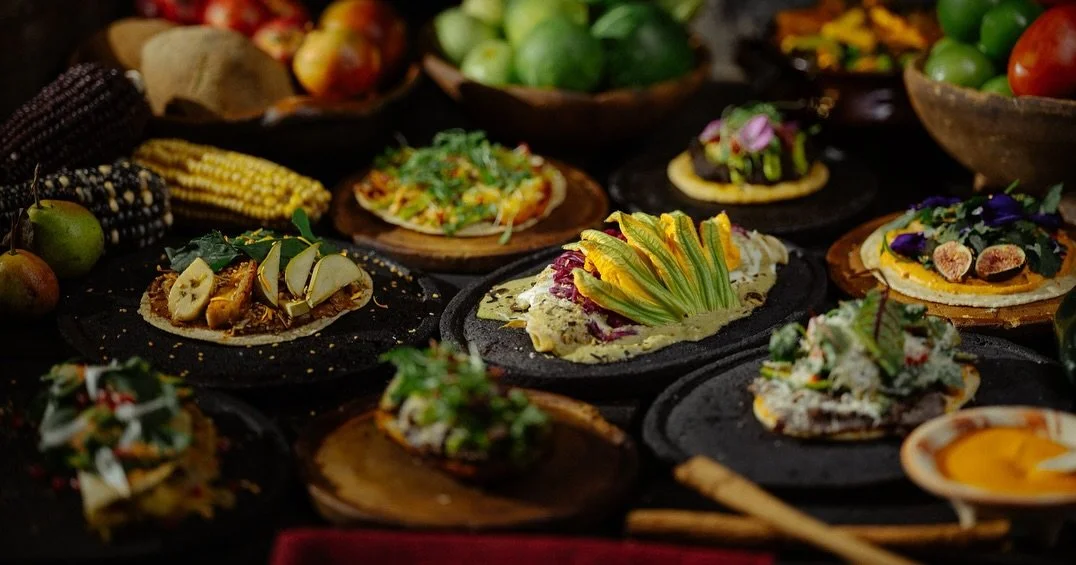 Assorted Mexican dishes and tacos served on black plates with various toppings and garnishes, along with fresh fruits and vegetables in the background.