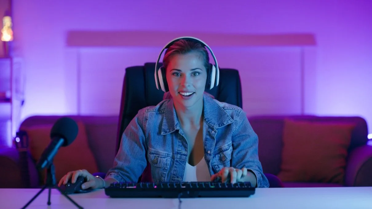 A young woman with blonde hair wearing headphones, sitting at a desk with a microphone, keyboard, and mouse, in a room with purple lighting, smiling at the camera.