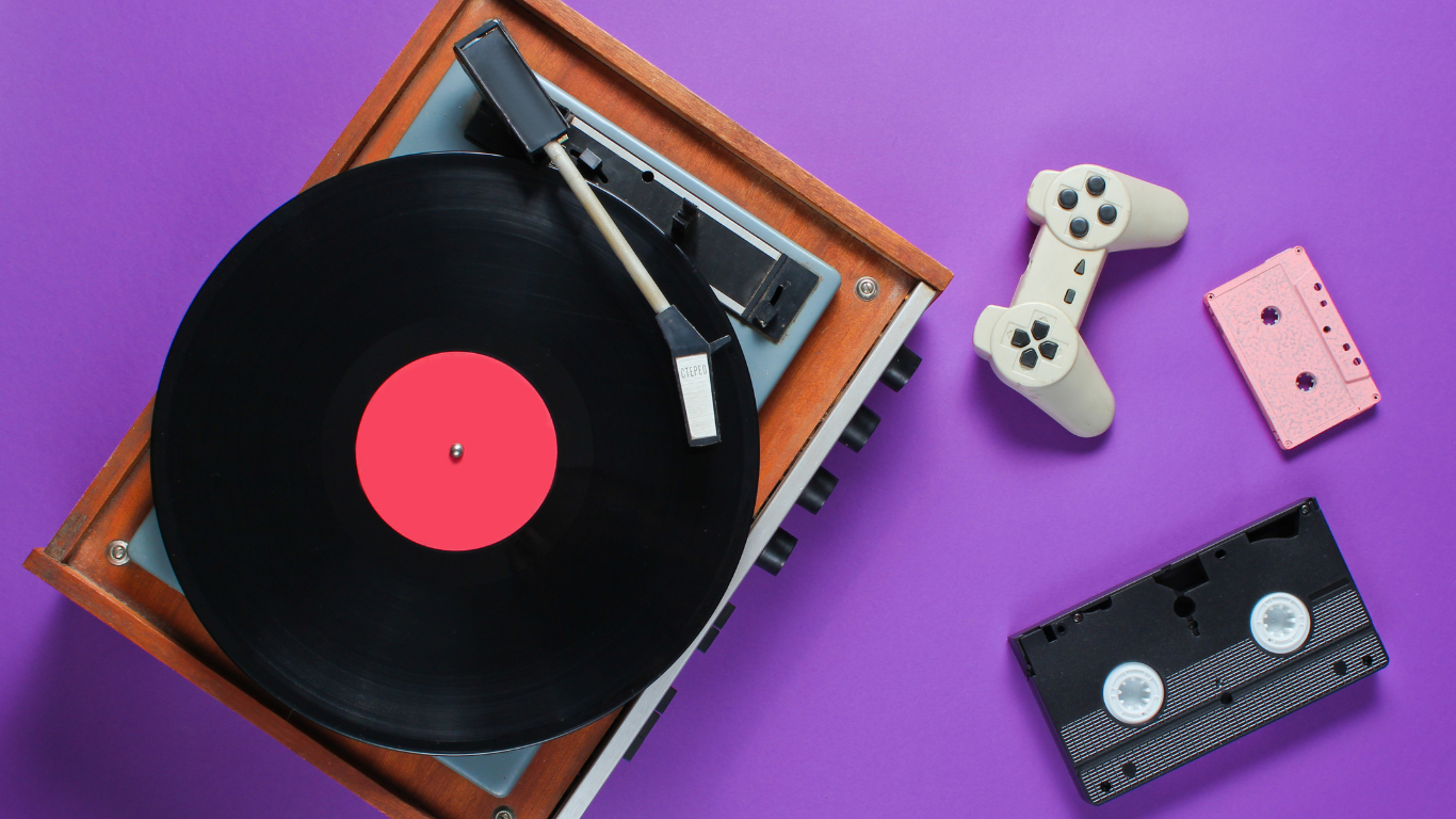 A vintage turntable playing a vinyl record, with a white game controller, a pink cassette tape, and a black cassette tape on a purple background.