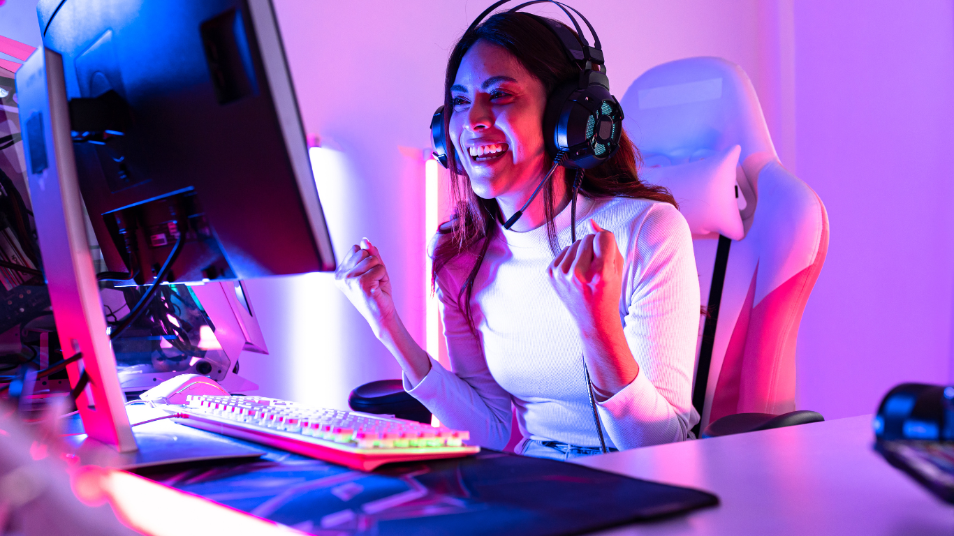 A woman sitting at a desk in front of a computer monitor, wearing a headset, smiling and celebrating with her fists clenched, in a colorful gaming setup with neon lights.