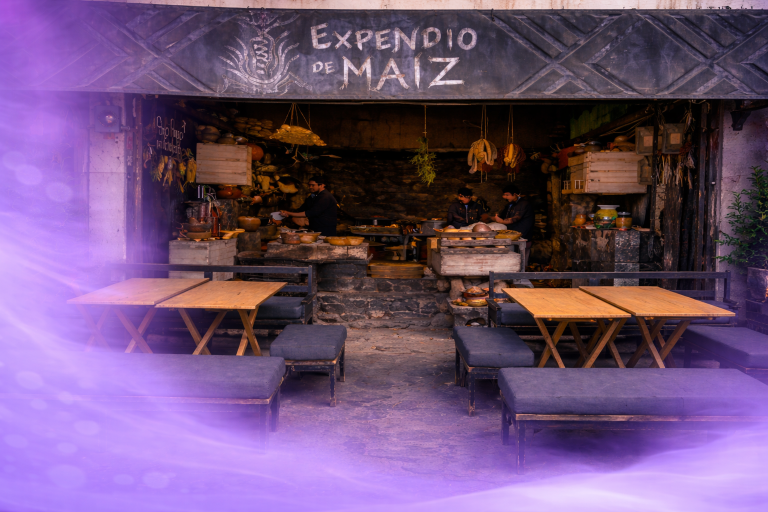 Open-air traditional Mexican restaurant called Expendio de Maíz with a rustic stone interior, wooden tables and benches, and chefs preparing food inside.