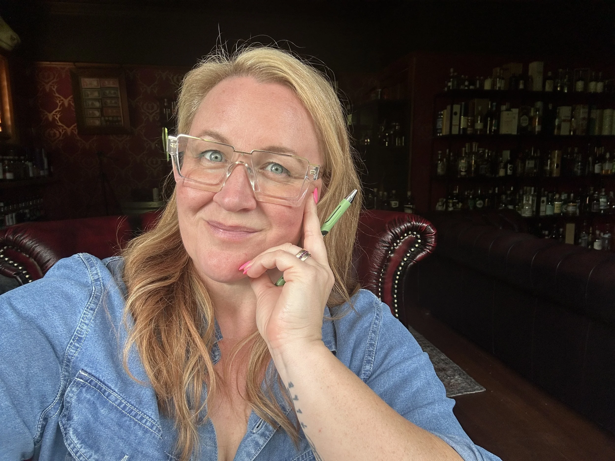 Tash McGill with long copper blonde hair wearing clear glasses and a denim shirt, smiling while holding a green pen, sitting on a red leather couch in a dimly lit room with shelves of bottles and dark floral wallpaper in the background.