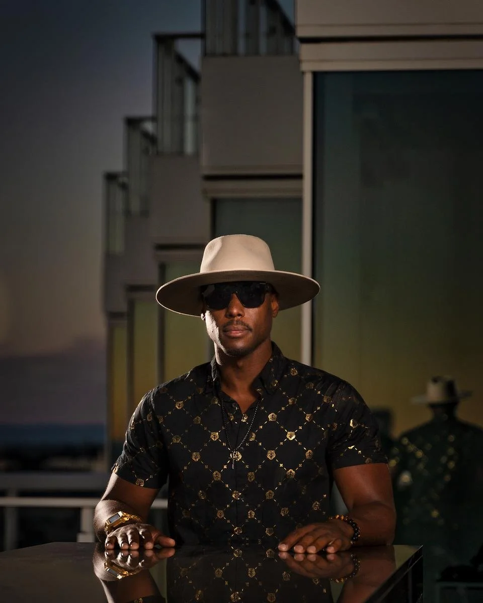 DJ NOÉL wearing a beige wide-brimmed hat, black sunglasses, a black shirt with a gold pattern, and jewelry, sitting at a reflective table in front of modern buildings during the evening or dusk.
