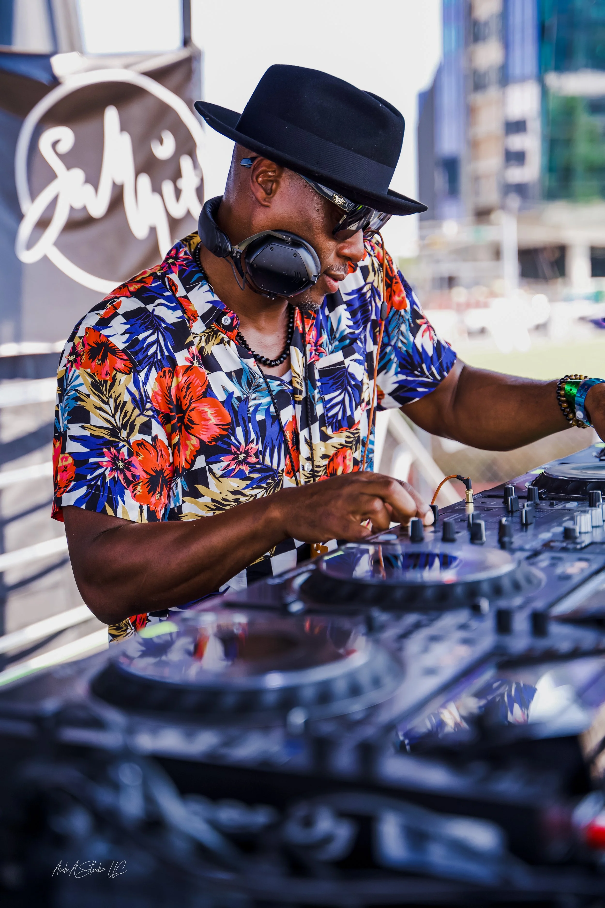 NOÉL wearing a black hat, sunglasses, and a colorful Hawaiian shirt, working at a DJ booth outside.
