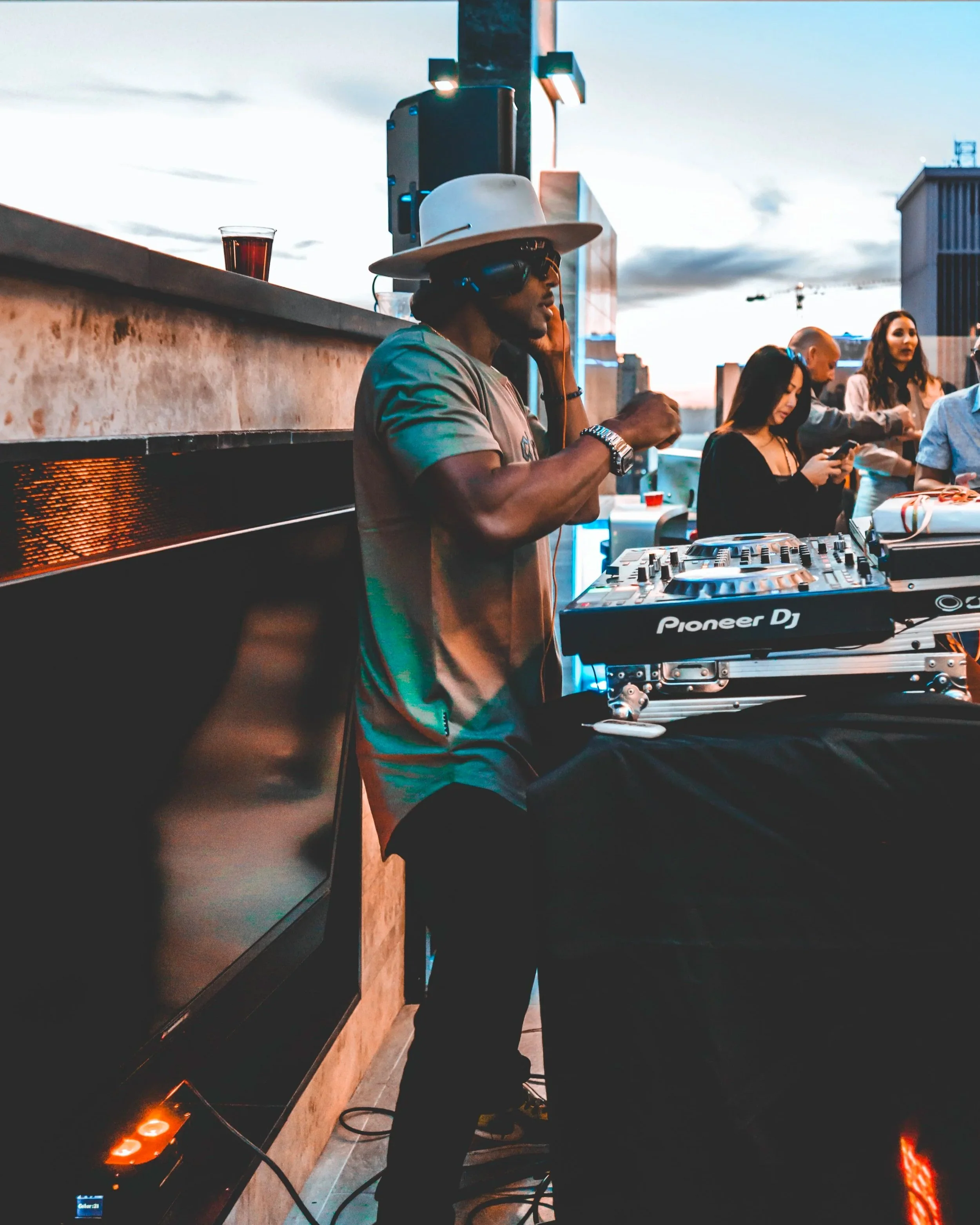 Artist DJ Producer NOÉL wearing a white hat and sunglasses, operating a Pioneer DJ setup on a rooftop during sunset, with a group of people in the background.