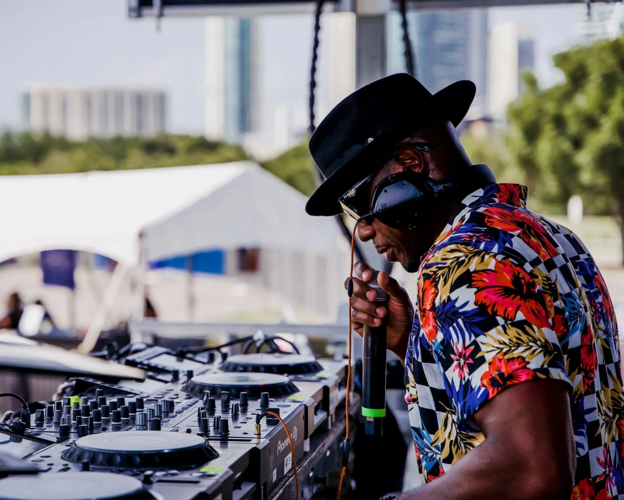 NOÉL wearing a floral shirt, black hat, and headphones, speaking into a microphone while operating DJ equipment outdoors with city buildings and trees in background.