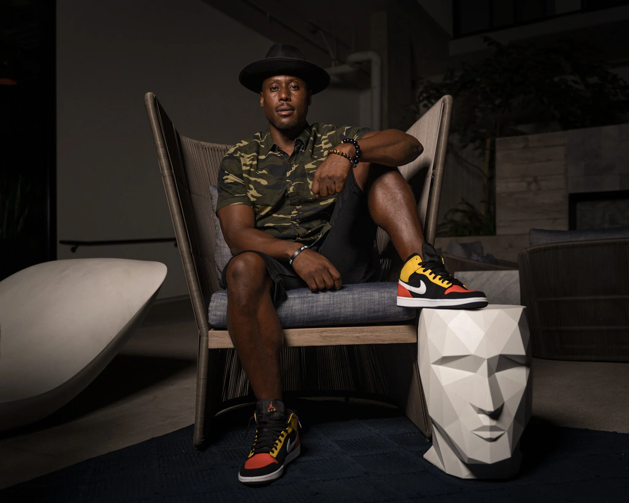 NOÉL wearing a camouflage shirt, black shorts, and colorful Nike sneakers sits in a modern chair indoors at night, with a white decorative face sculpture on the floor beside him.