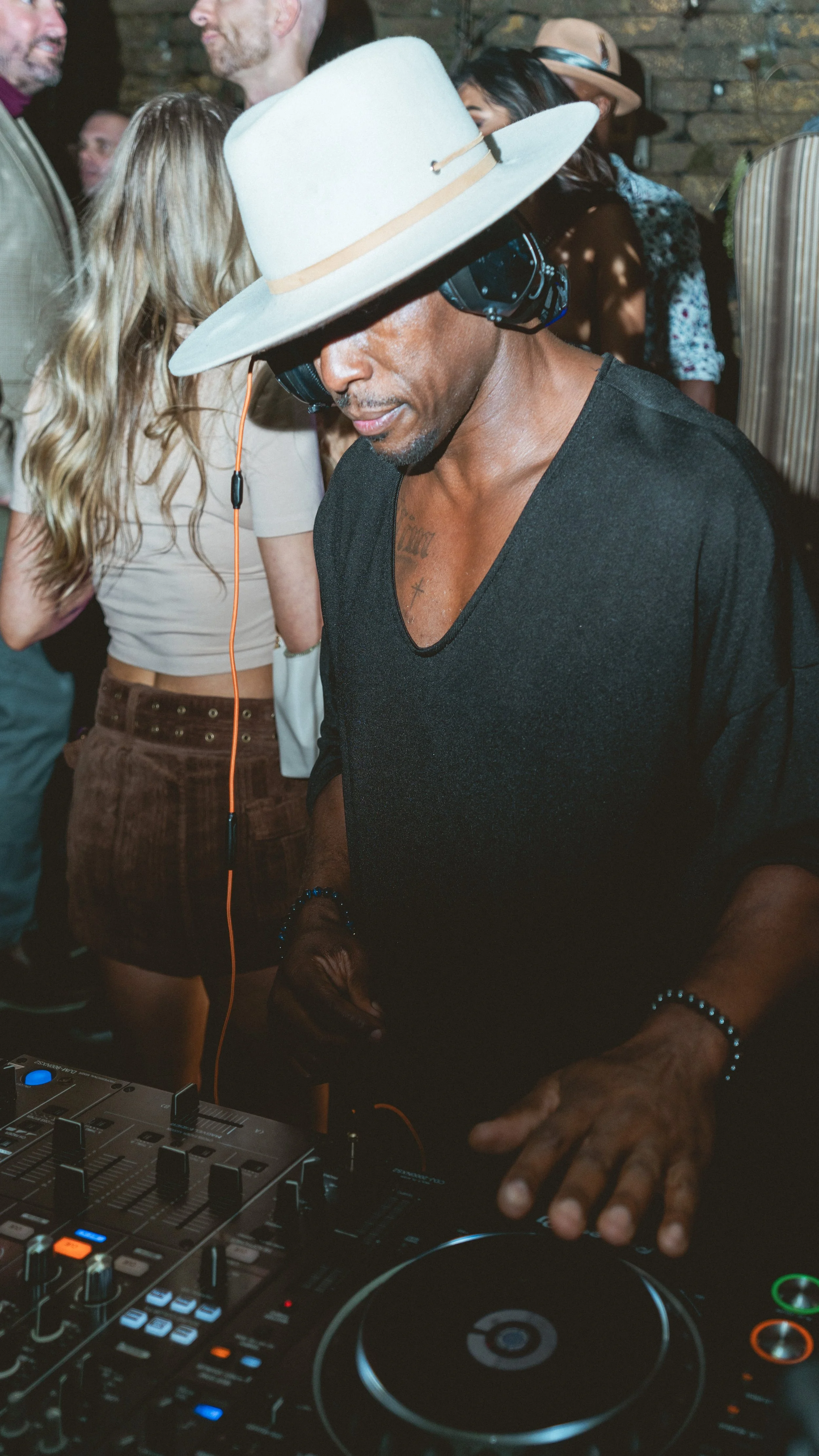 Artist DJ Producer NOÉL wearing a large white hat, headphones, and a black shirt is mixing music at a party with a crowd dancing behind him.