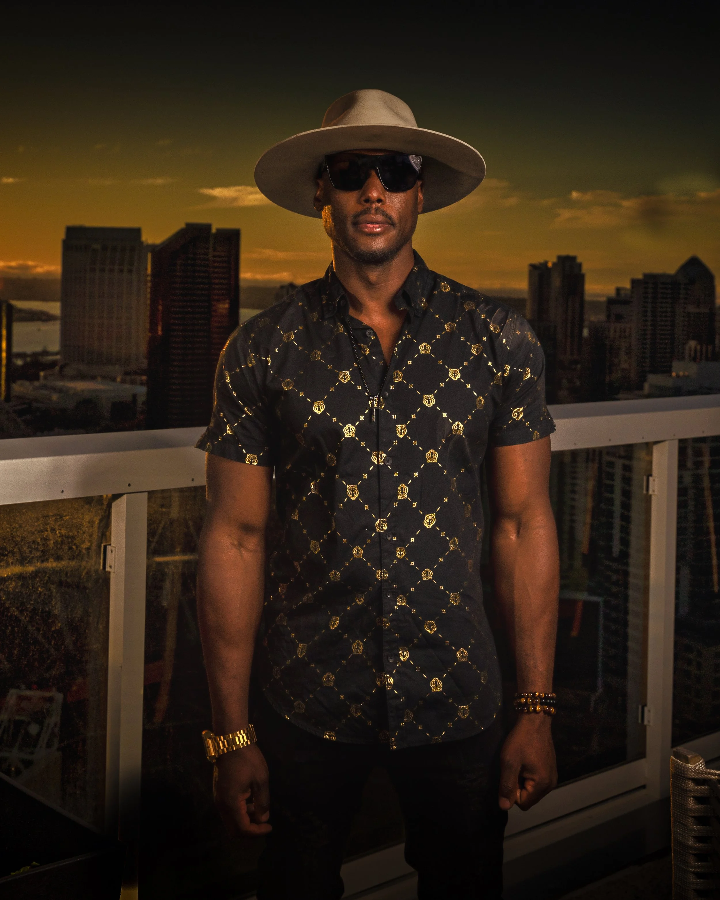 NOÉL standing on a balcony at sunset, wearing sunglasses, a wide-brimmed hat, a patterned black shirt with gold accents, and gold accessories.