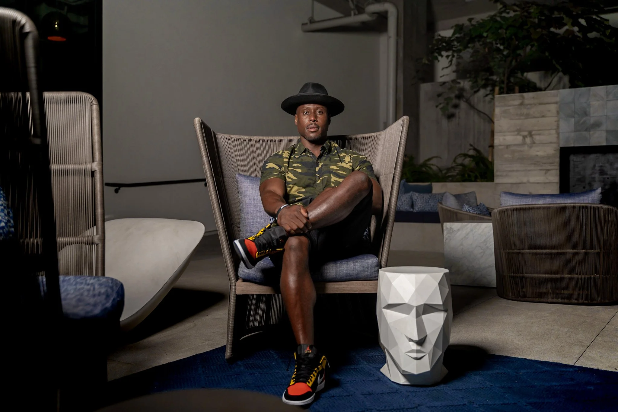 Artist DJ Producer NOÉL wearing a black hat, camo shirt, and sneakers sitting cross-legged on a modern outdoor chair in a contemporary patio setting with decorative furniture and a geometric face-shaped planter.