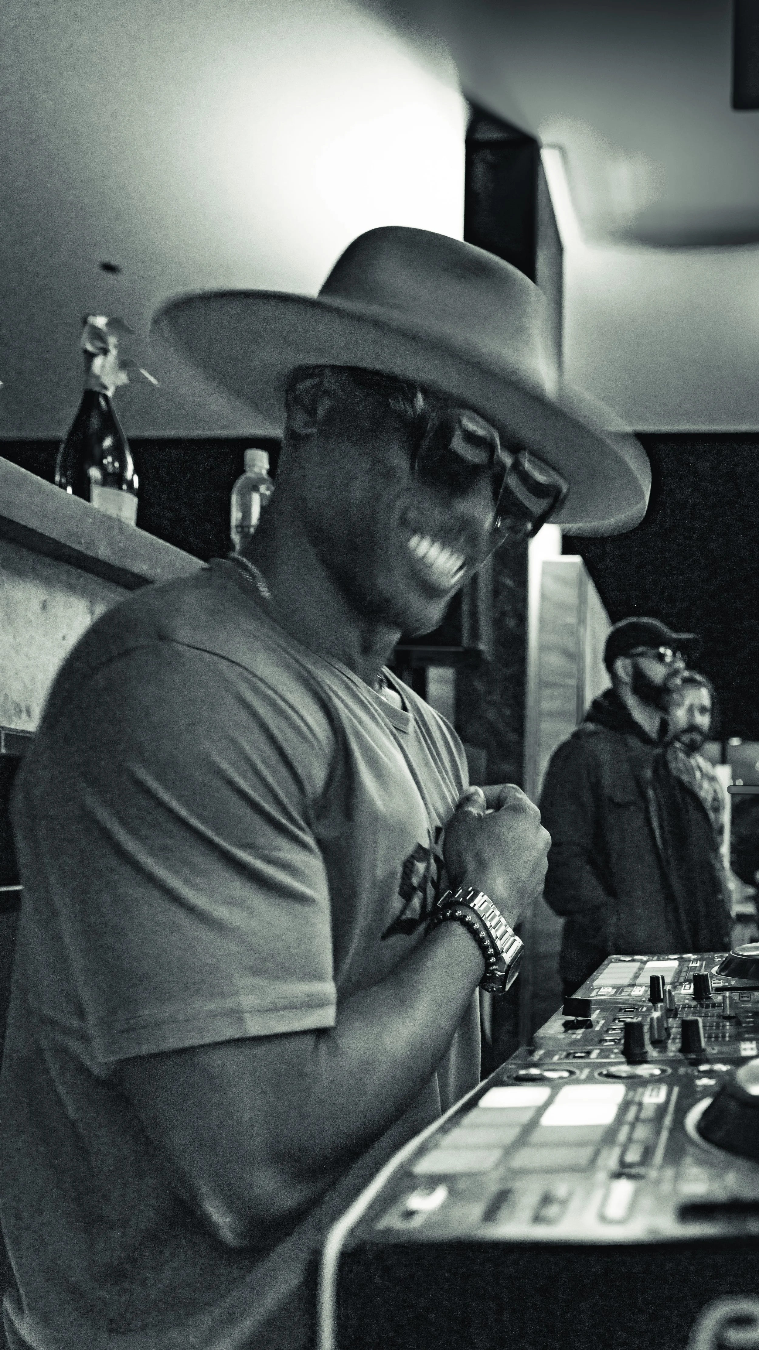 Artist DJ Producer NOÉL glasses, a large hat, and a T-shirt, smiling and posing with at a DJ booth, with two other men in the background, in a dimly lit indoor setting.
