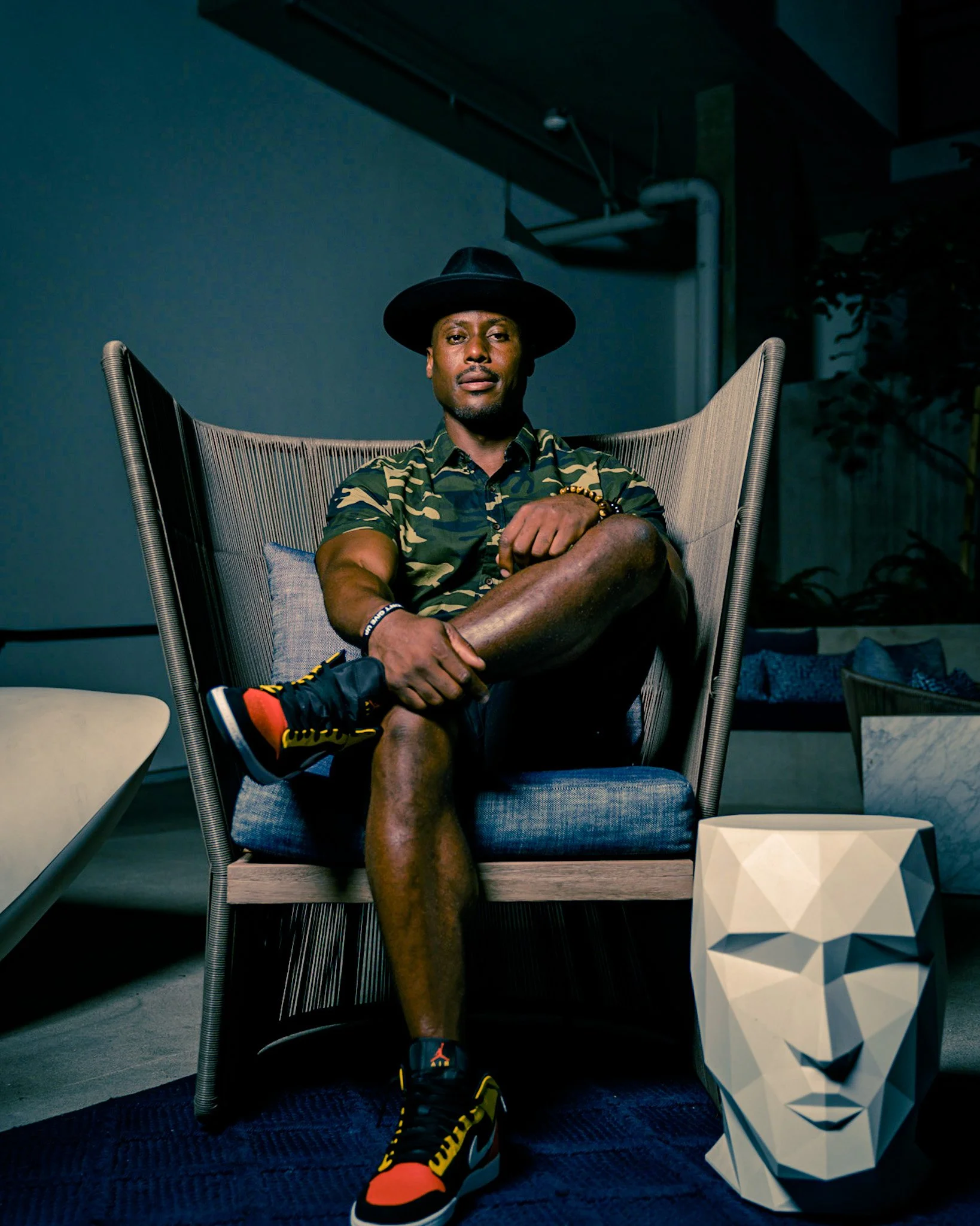 Artist DJ Producer NOÉL in a modern chair in a dimly lit room, wearing a black hat, camouflage shirt, black shorts, and colorful sneakers, with a geometric human face sculpture on the floor nearby.