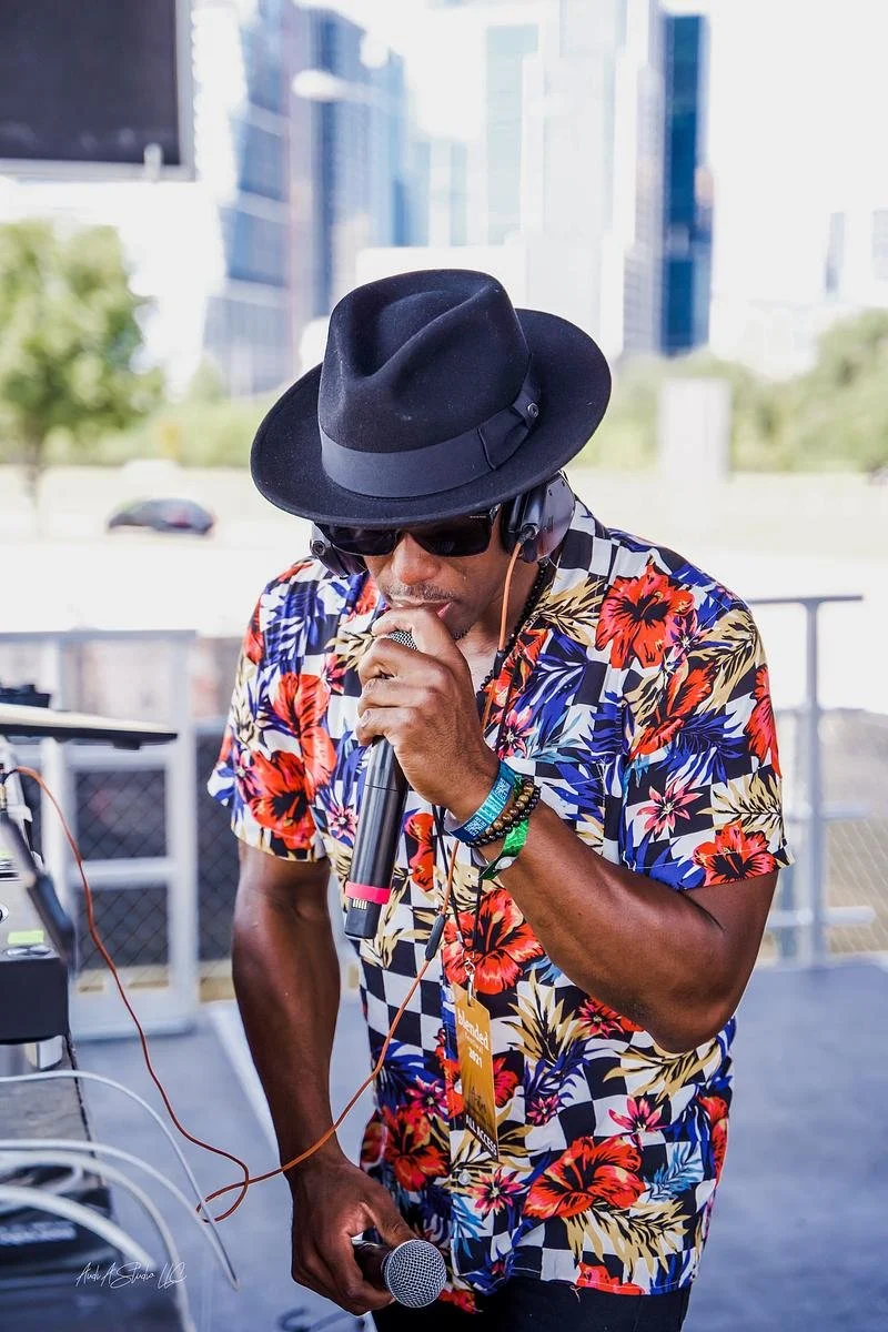Artist NOÉL wearing a black fedora hat, sunglasses, and a colorful floral shirt is performing with a microphone, surrounded by DJ equipment outdoors with city buildings in the background.