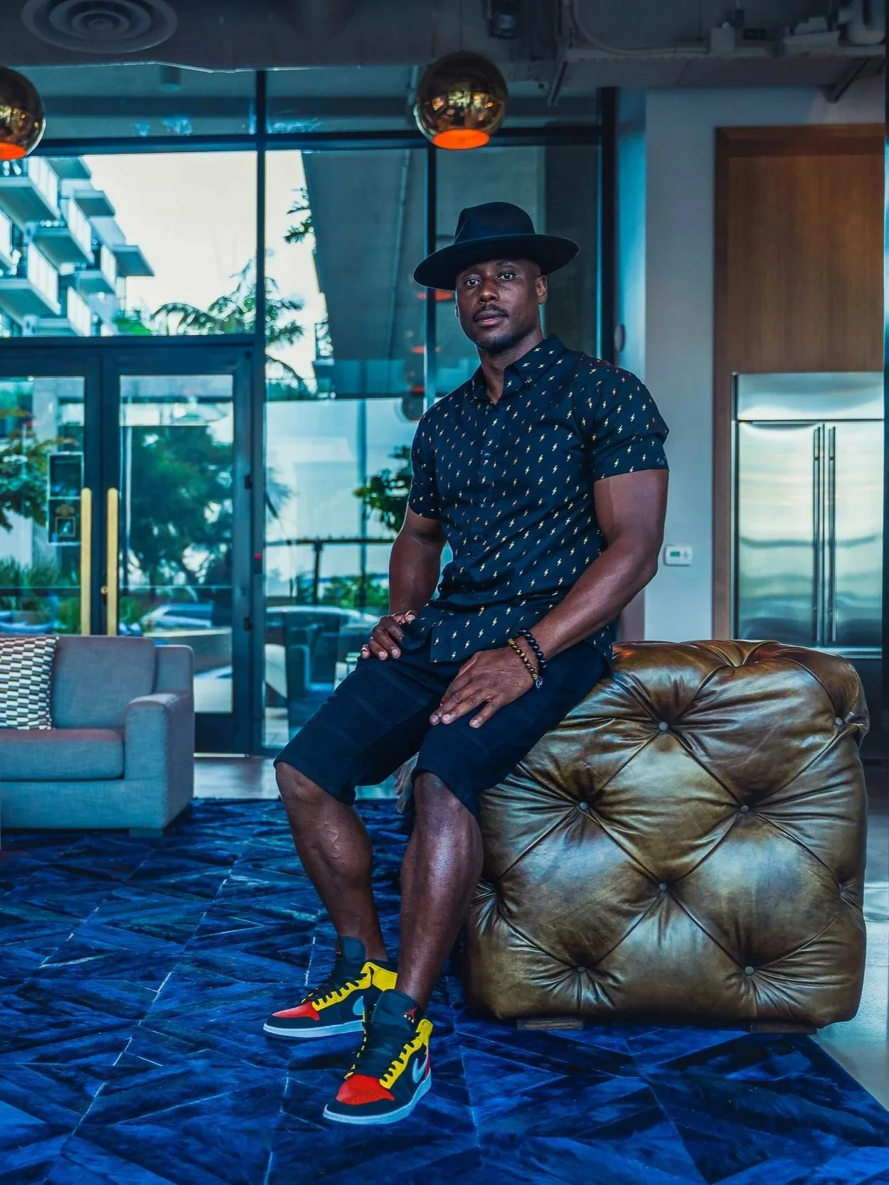 Artist DJ Producer NOÉL sitting on a large, tufted leather ottoman inside a modern building with glass doors and windows, wearing a black hat, a patterned short-sleeve shirt, shorts, and colorful sneakers.