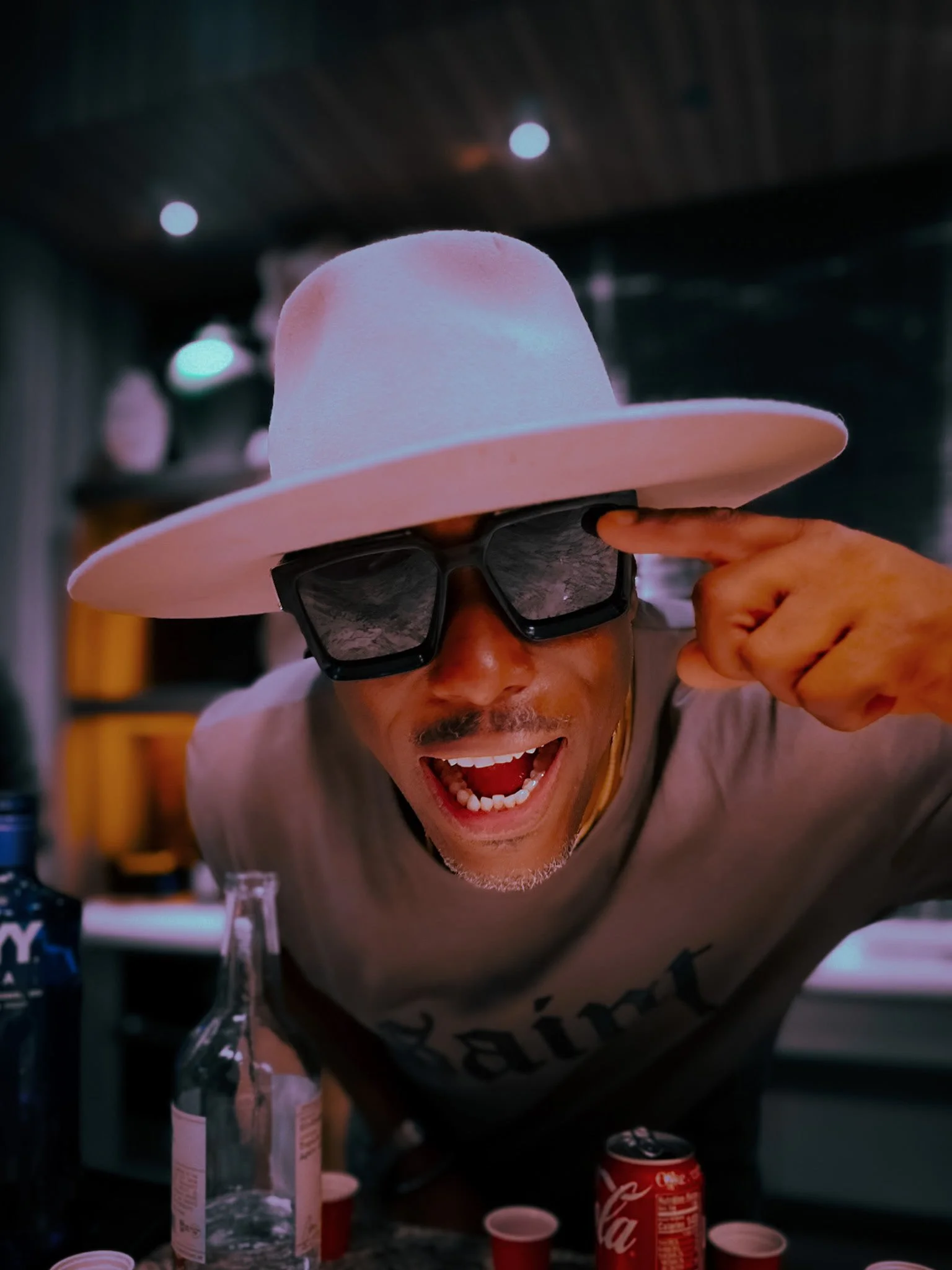 Artist DJ Producer NOÉL wearing a light beige wide-brim hat and dark sunglasses is smiling and pointing to his head with his index finger. At an afters