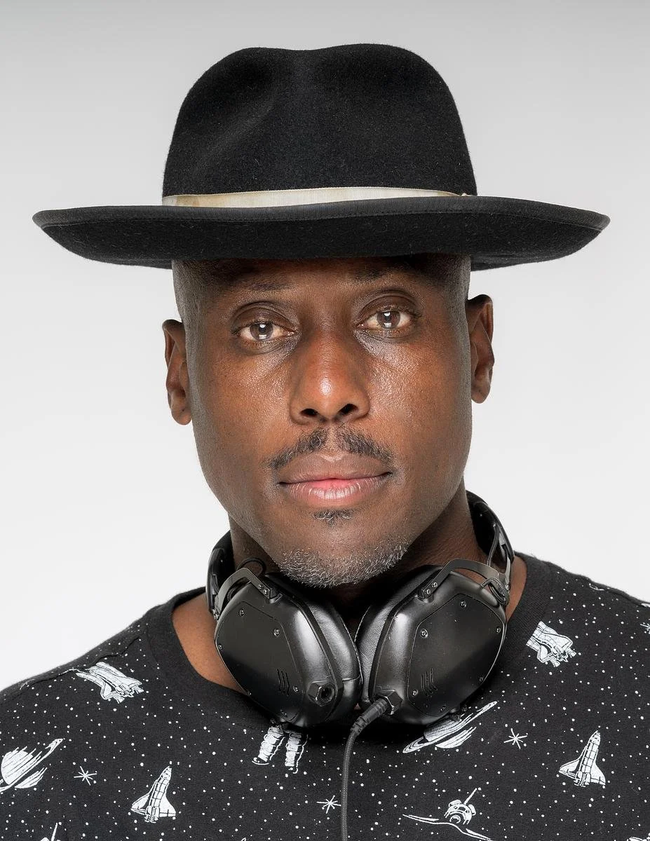 A man wearing a black hat with a beige band, black headphones around his neck, and a black shirt with white space-themed illustrations.