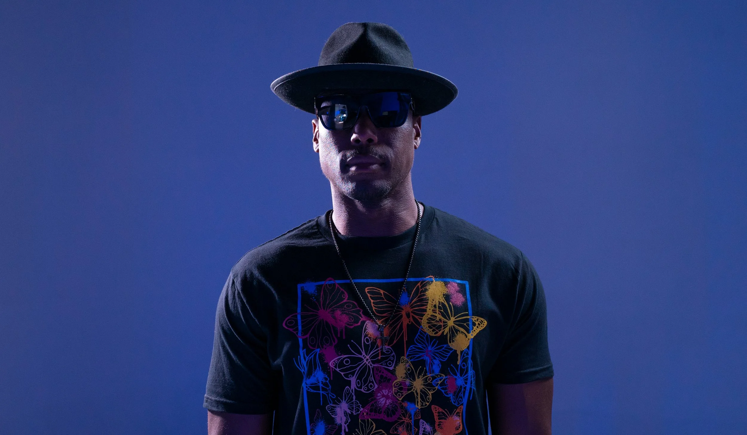 NOÉL wearing a dark wide-brimmed hat, sunglasses, a black graphic T-shirt with colorful butterfly designs, and a necklace stands against a plain blue background.