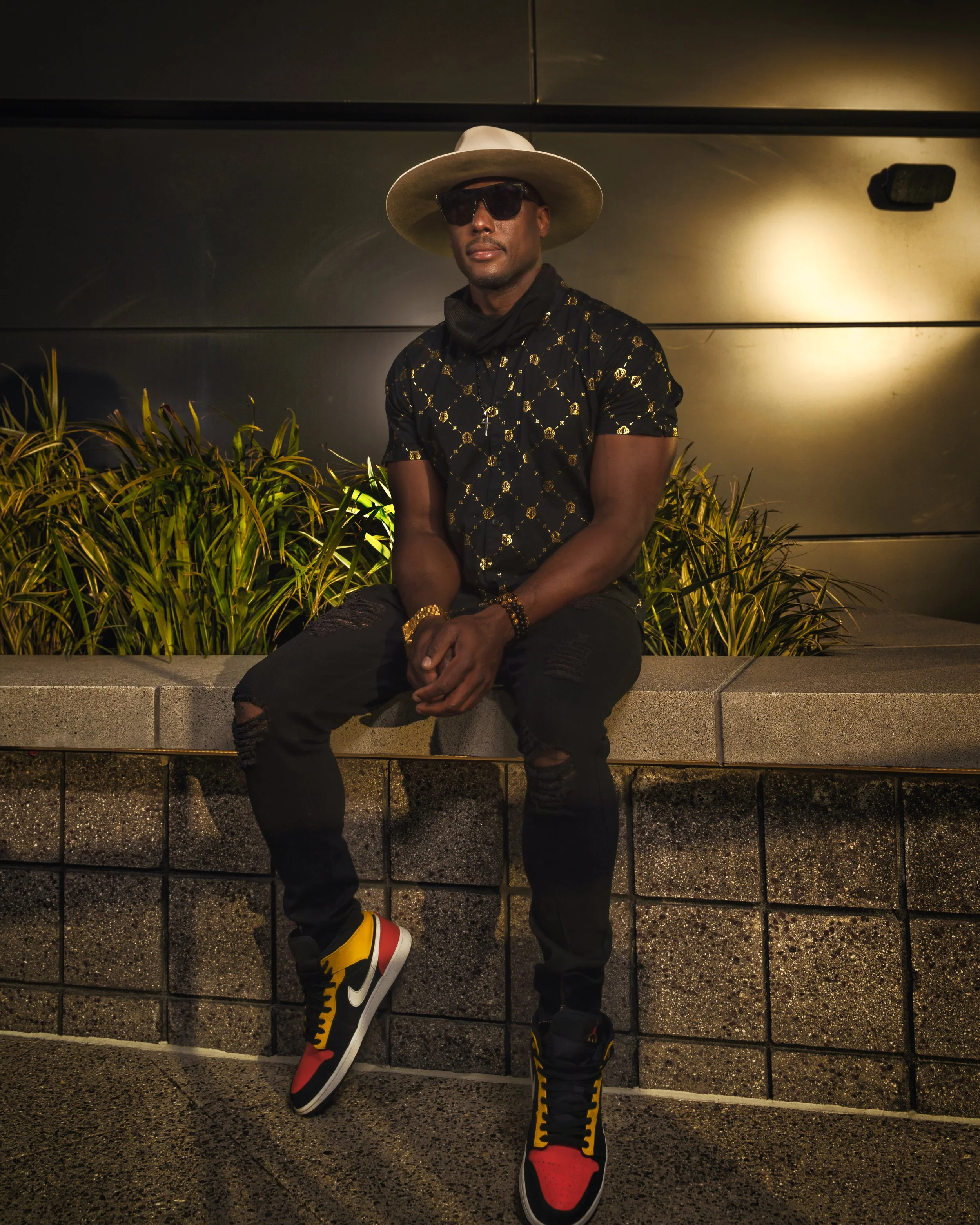 NOÉL sitting on a concrete ledge at night, wearing a wide-brimmed hat, sunglasses, a black patterned shirt, distressed black jeans, and colorful Jordans. The background features plants and a dark wall with warm lighting.