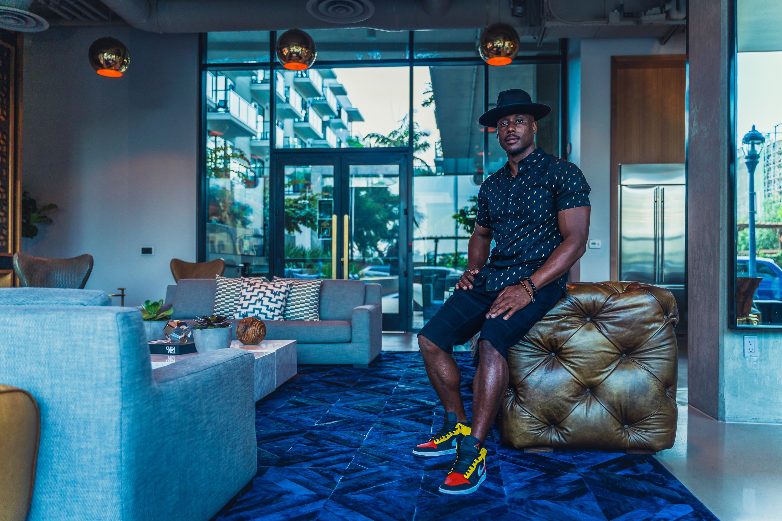 black hat, patterned shirt, black shorts, and colorful sneakers sits on a leather ottoman in a modern hotel lobby with comfortable chairs, decorative pillows, a coffee table with plants and decor, and large glass doors showing an outside view.