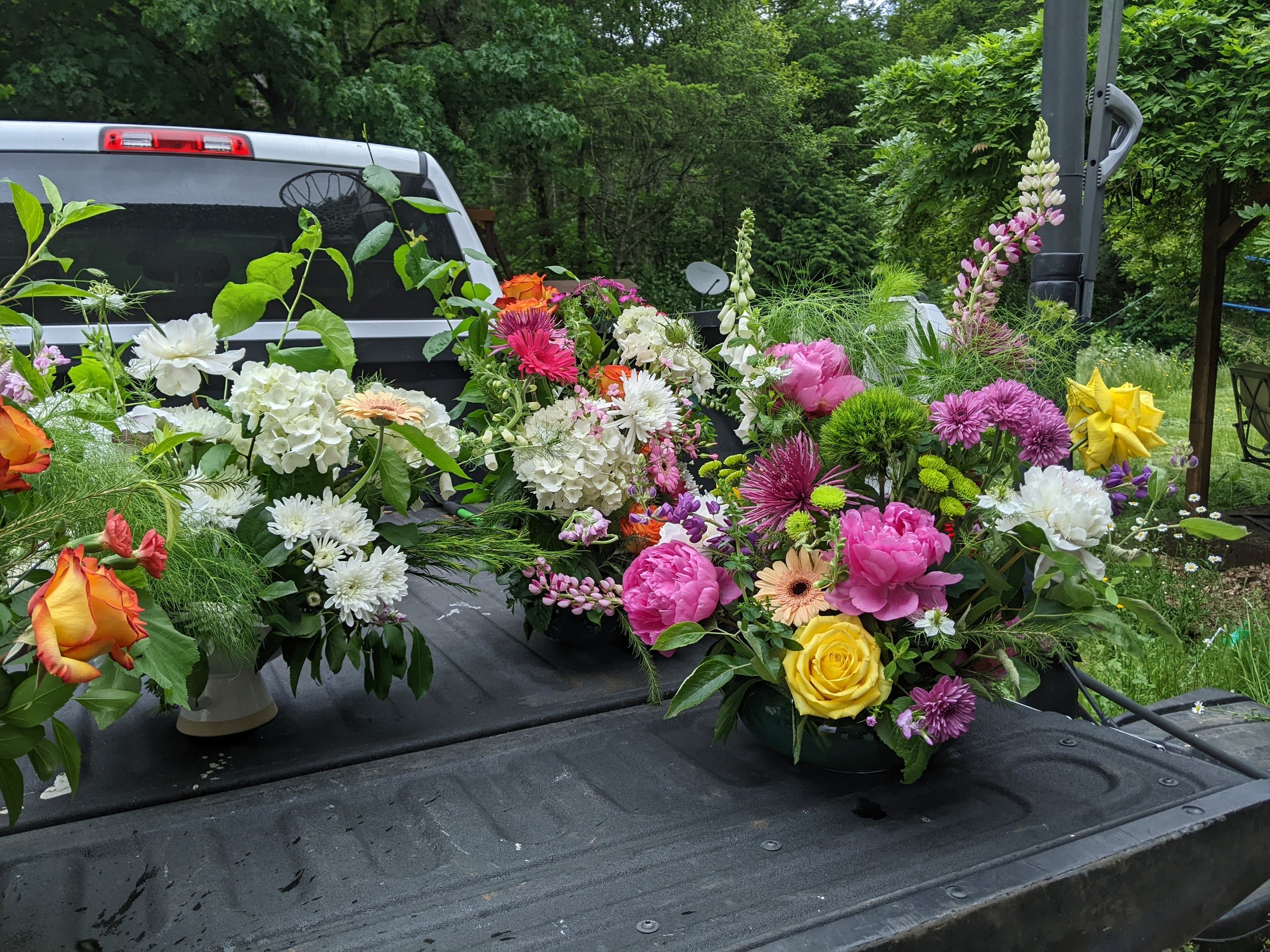 Multiple colorful flower arrangements on the back of a black truck bed, with a green rural landscape in the background.