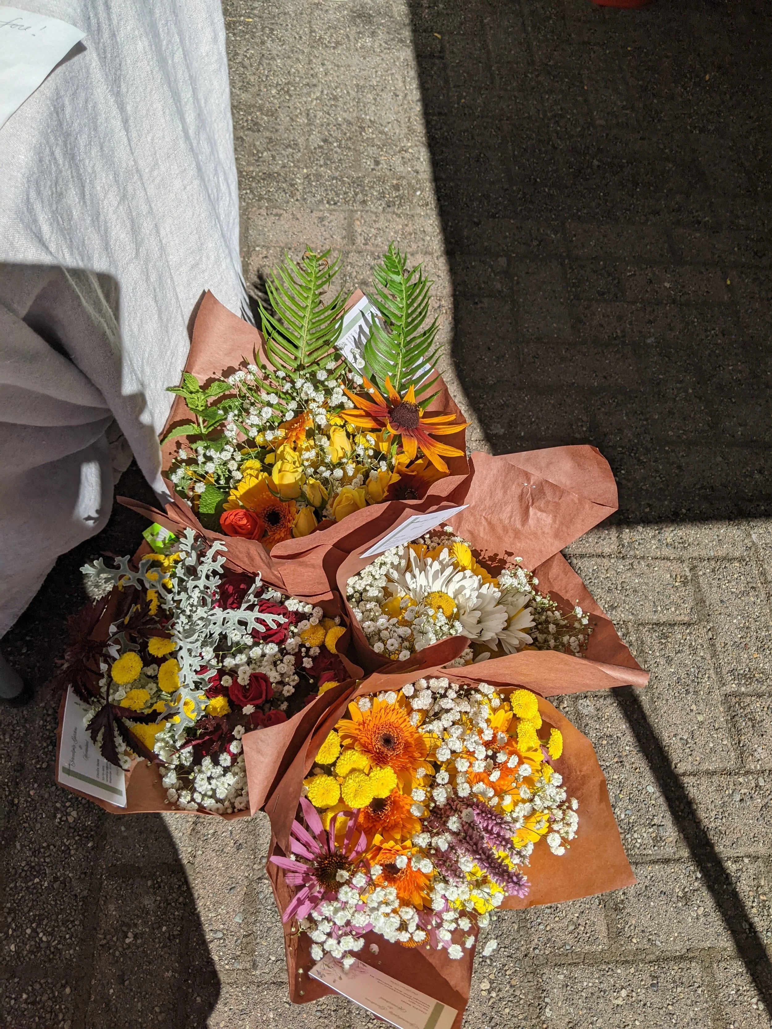 Multiple bouquets of colorful flowers, including daisies, chrysanthemums, and other mixed blooms, wrapped in brown paper, lying on a concrete sidewalk.