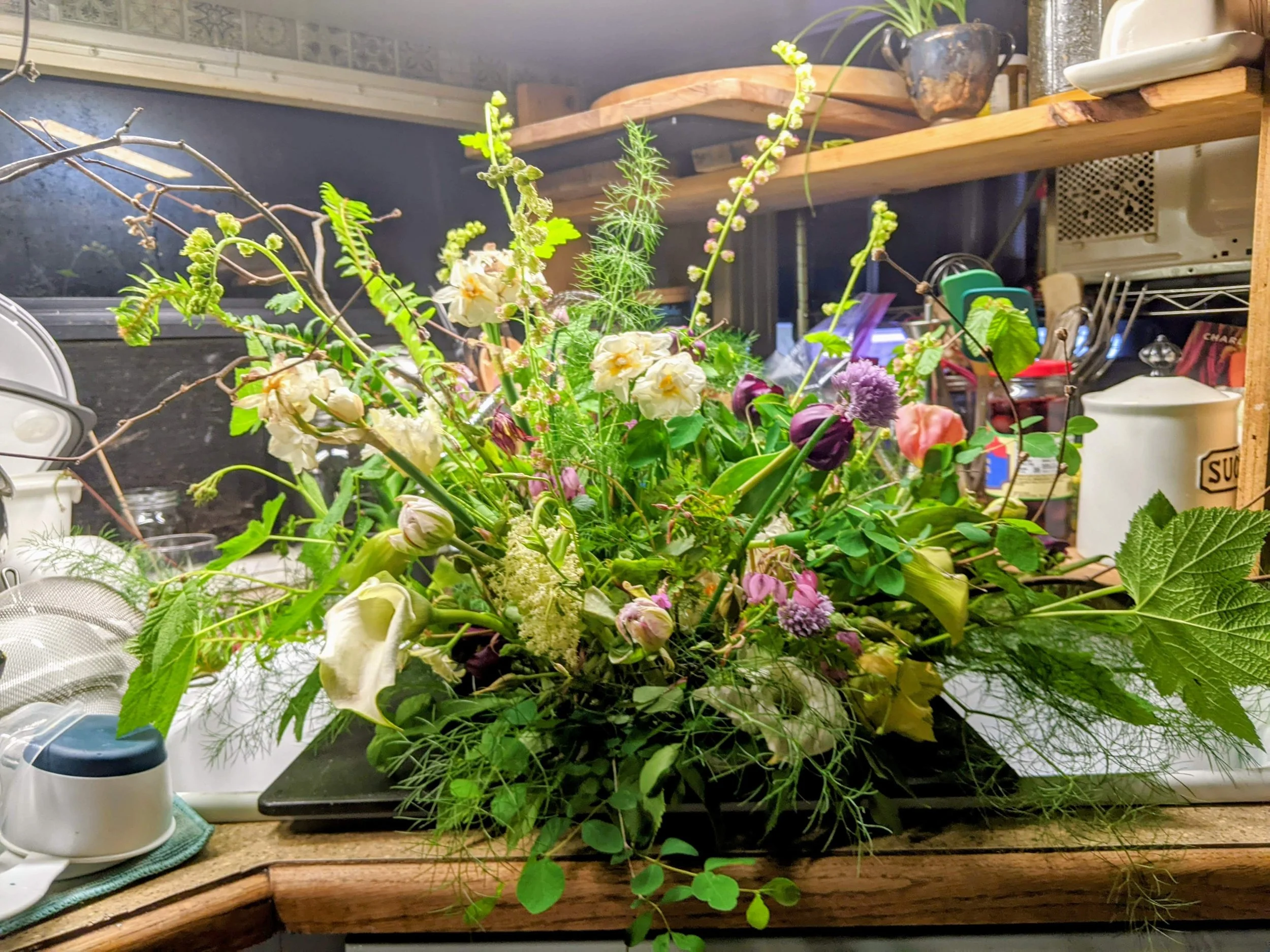 A large, colorful floral arrangement with white, purple, pink, and green flowers and leaves on a kitchen countertop with various kitchen items in the background.