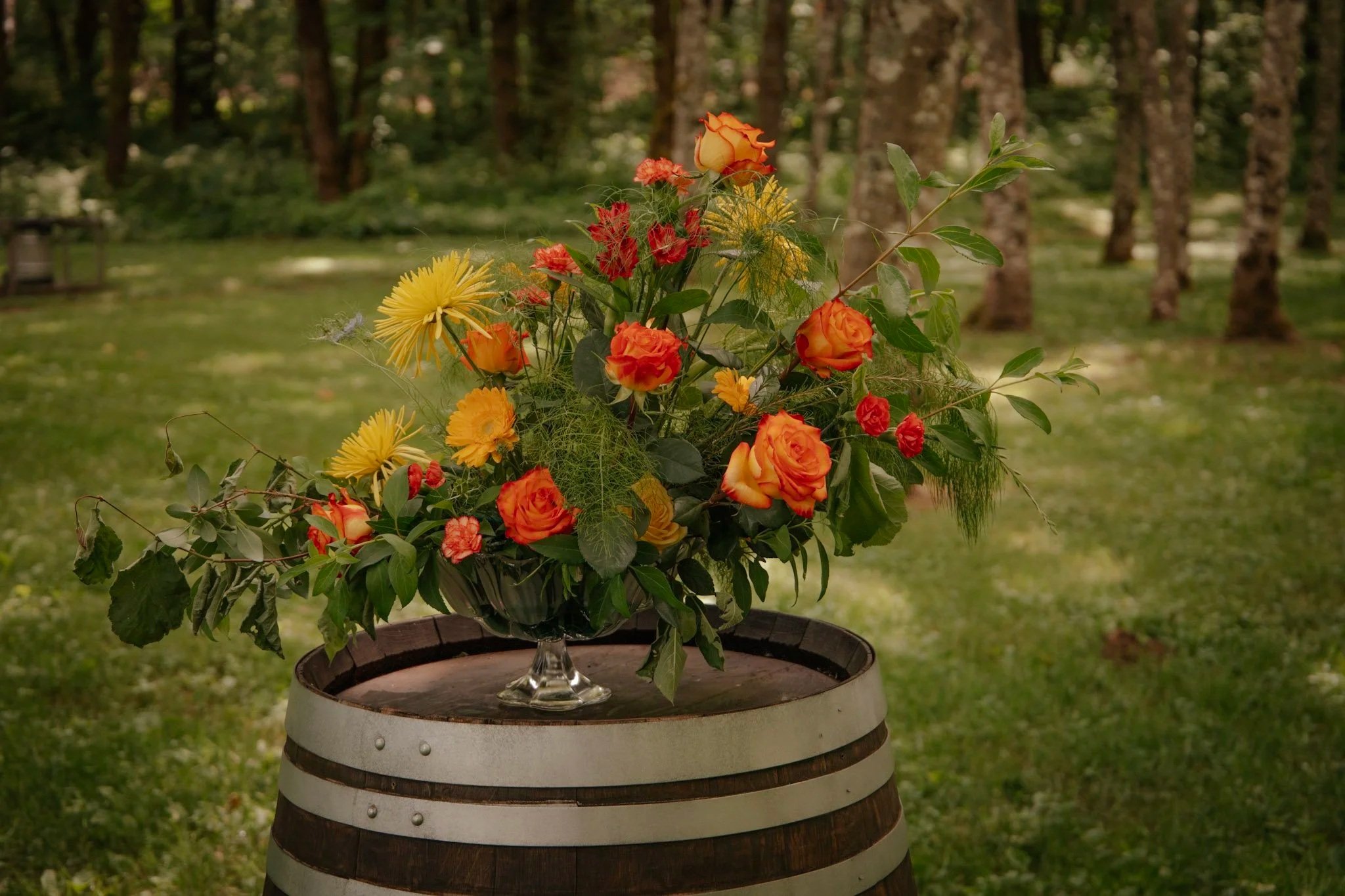 A colorful bouquet of orange and yellow flowers in a glass vase on top of a wooden barrel outdoors in a grassy area.